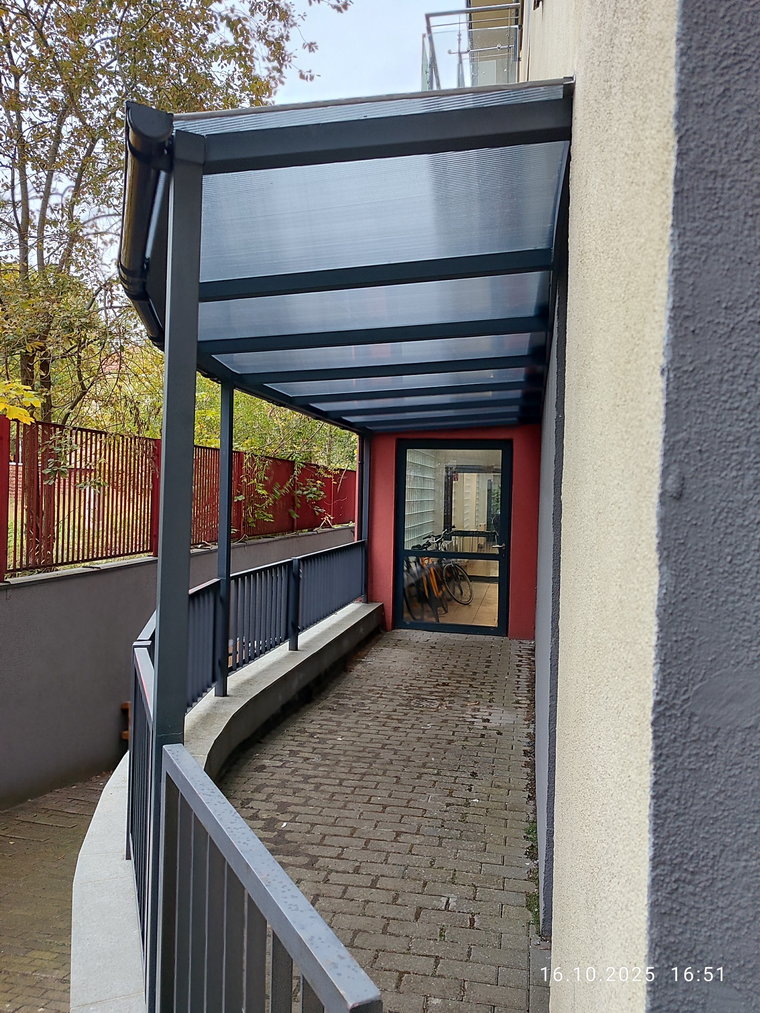 Metalowa pergola z przezroczystym zadaszeniem nad wejściem do budynku, widok z boku, z rowerami w tle. Szara konstrukcja, ceglana ścieżka, jesienne drzewa w tle.