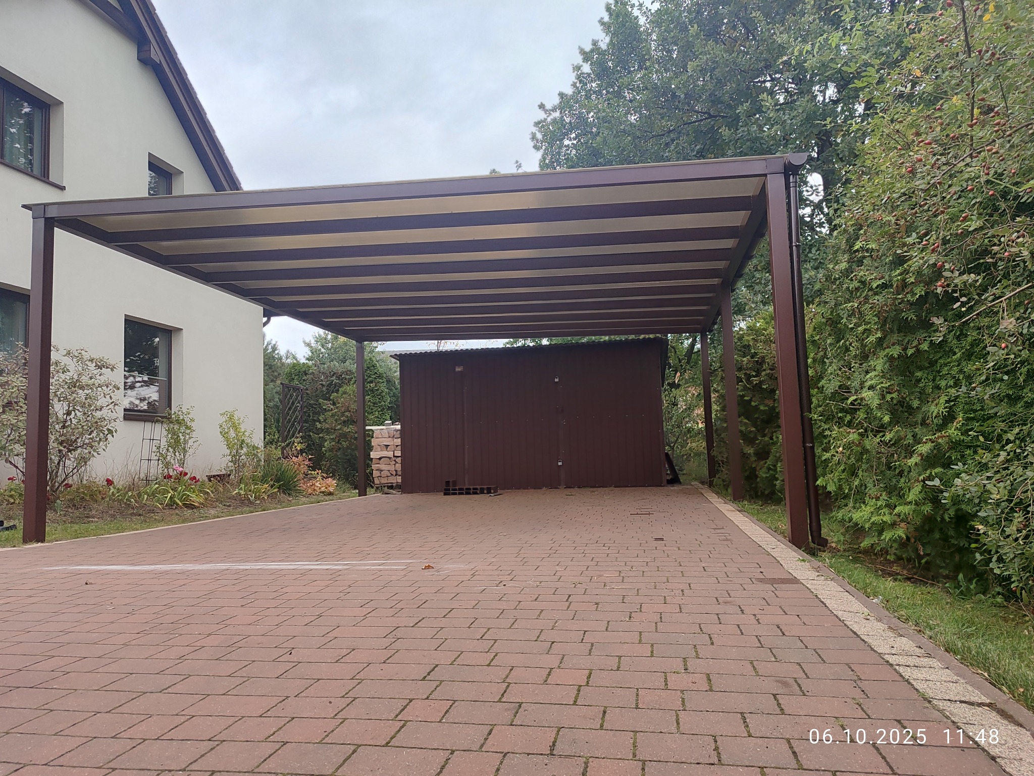 Nowoczesna, brązowa pergola metalowa z beżowymi pasami zadaszenia, przylegająca do domu z kostki brukowej. Widok na ogród i mały budynek gospodarczy w tle.