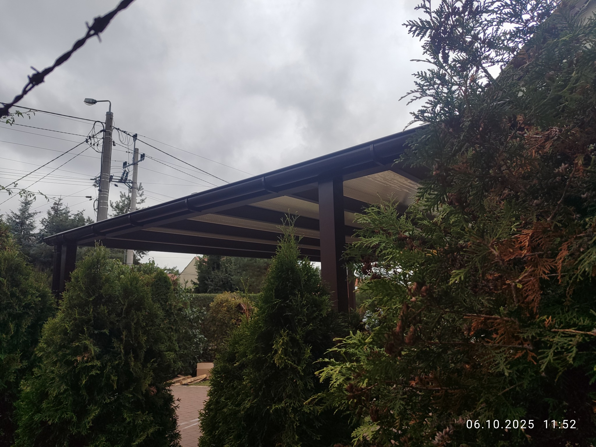 Nowoczesna, brązowa pergola z jasnym zadaszeniem, otoczona zielonymi tujami. Widok z dołu, pochmurne niebo w tle. Elementy infrastruktury miejskiej widoczne w tle.