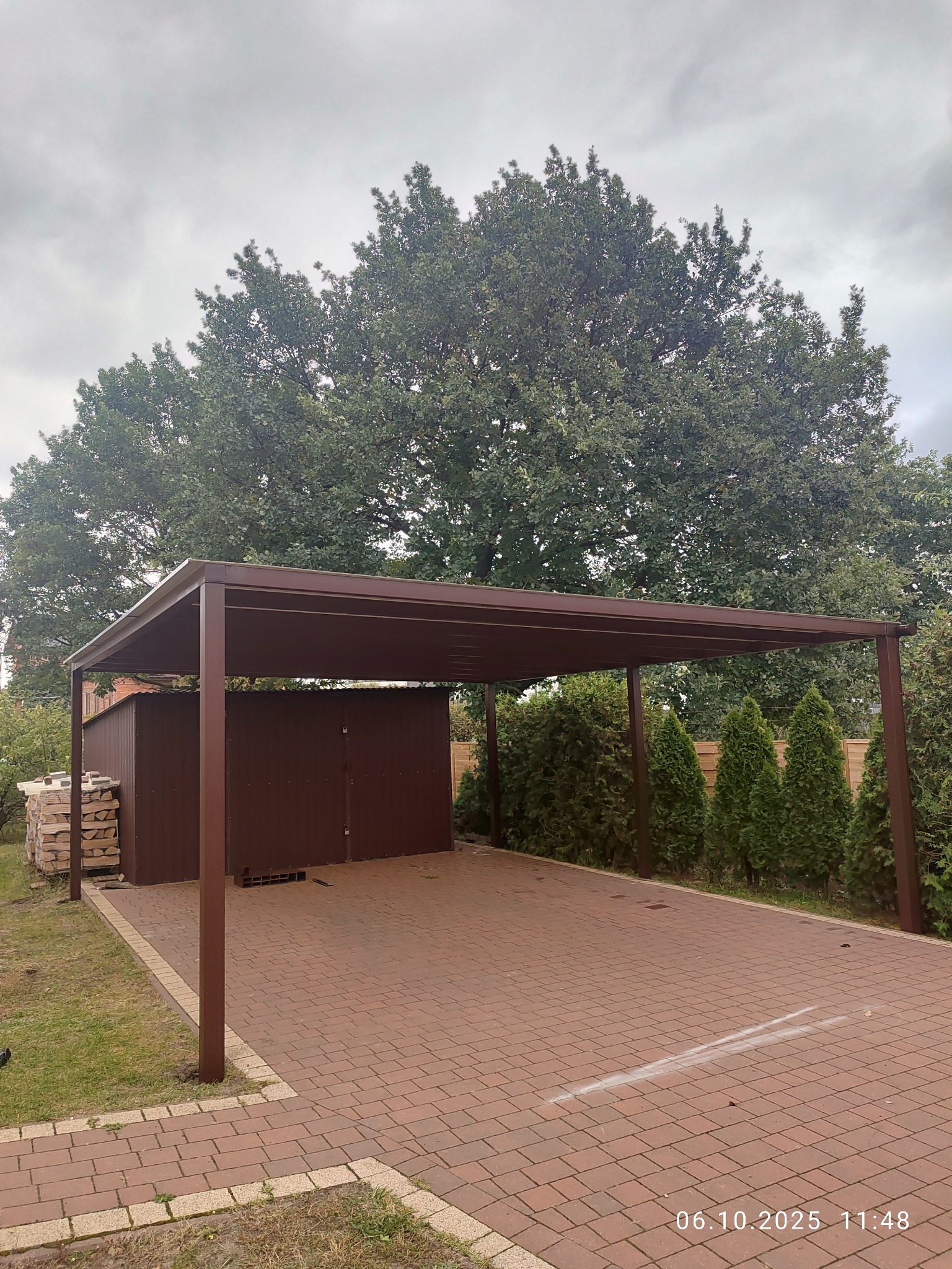 Brązowa, metalowa pergola na posesji z kostki brukowej, z widocznym składem drewna i zielenią w tle, sfotografowana w pochmurny dzień.