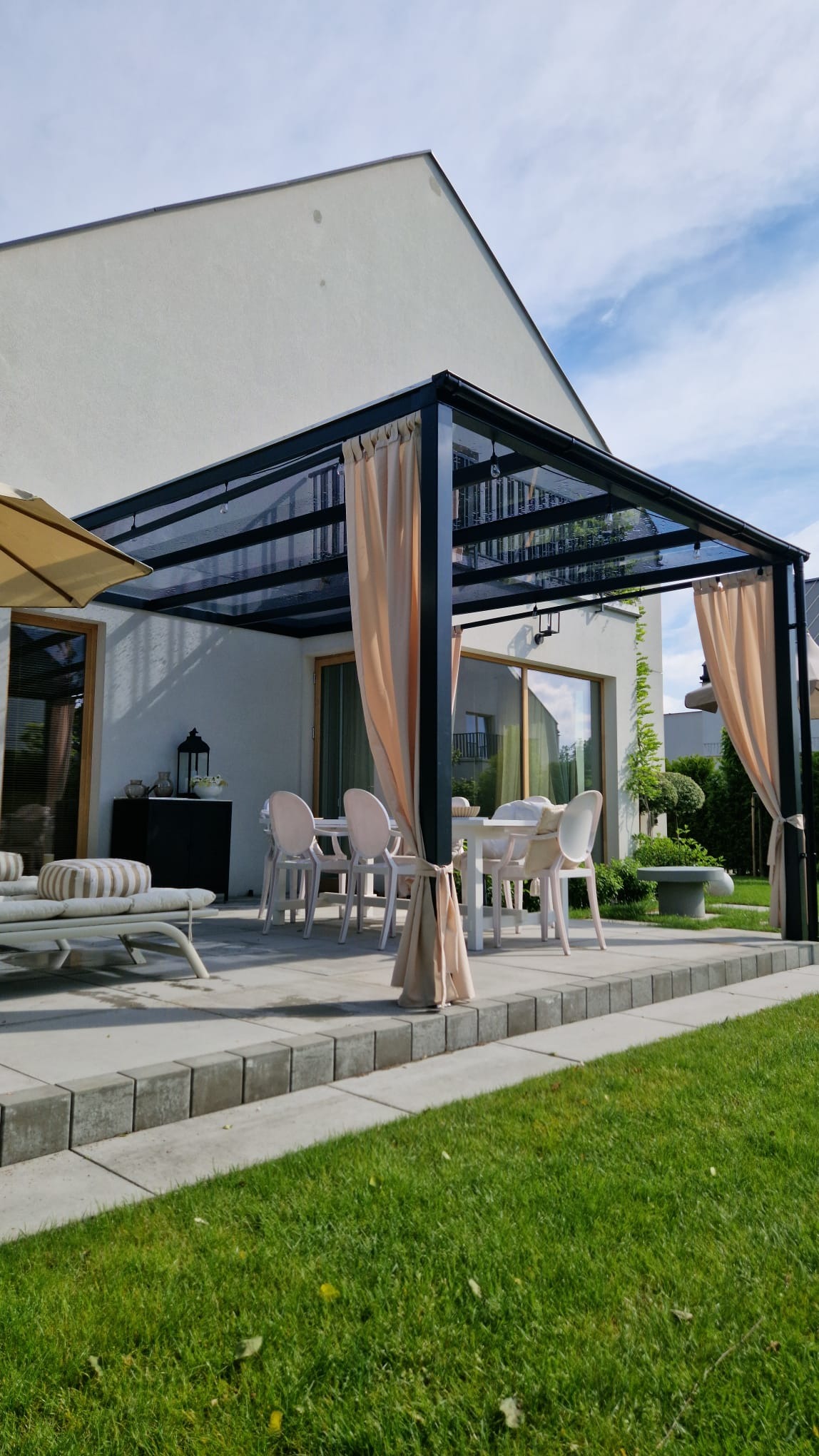 Nowoczesna, czarna pergola metalowa z przezroczystym dachem i beżowymi zasłonami, ustawiona na tarasie z jasnych płyt, z widokiem na zadbany trawnik i dom.