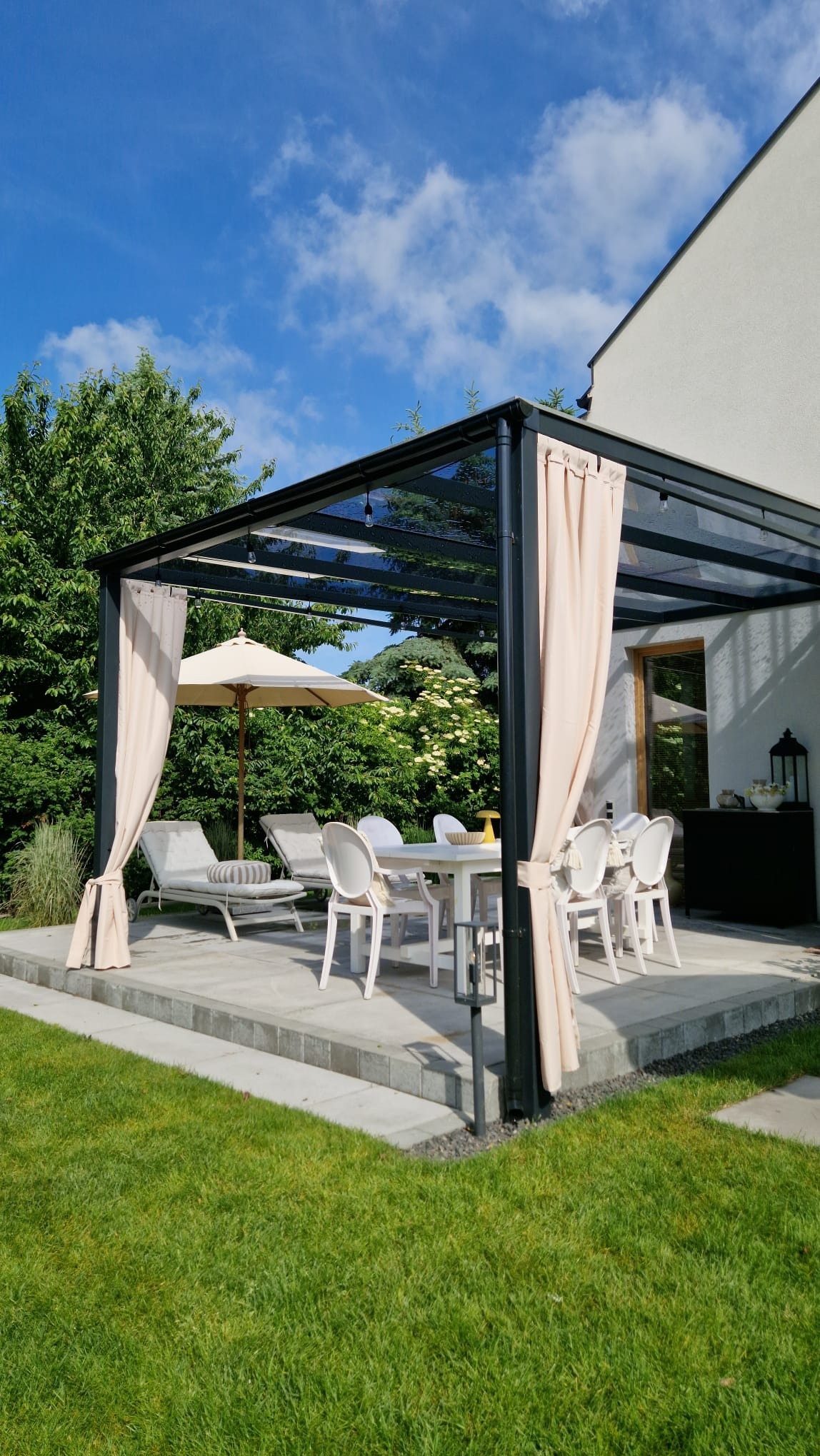 Nowoczesna, metalowa pergola z zasłonami w beżowym odcieniu, oświetleniem i meblami ogrodowymi na tarasie. Widok na zadbany ogród i błękitne niebo.