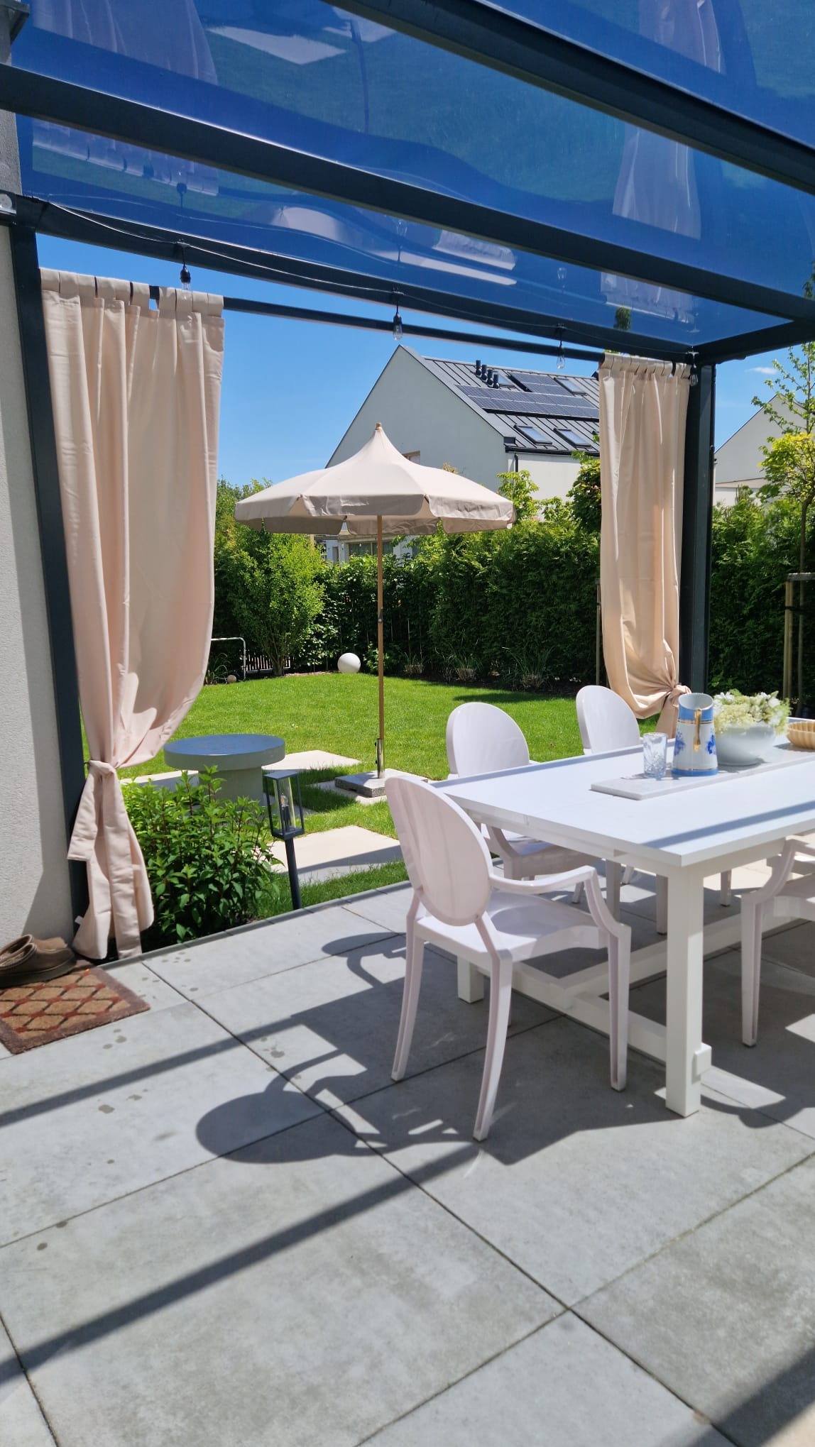 Metalowa pergola z przezroczystym dachem, beżowymi zasłonami i białymi meblami ogrodowymi na tarasie. W tle trawnik z parasolem i dom z panelami słonecznymi.