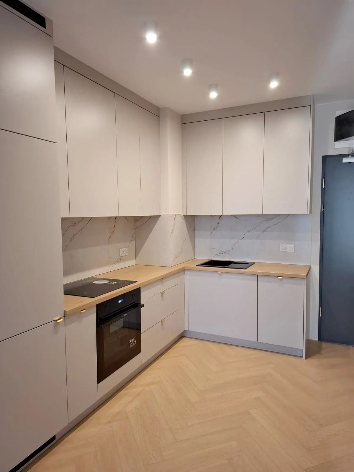 Jasna kuchnia w kształcie litery L z drewnianym blatem i marmurowym backsplashiem. Minimalistyczne fronty szafek z delikatnymi, złotymi uchwytami. Parkiet w jodełkę na podłodze.