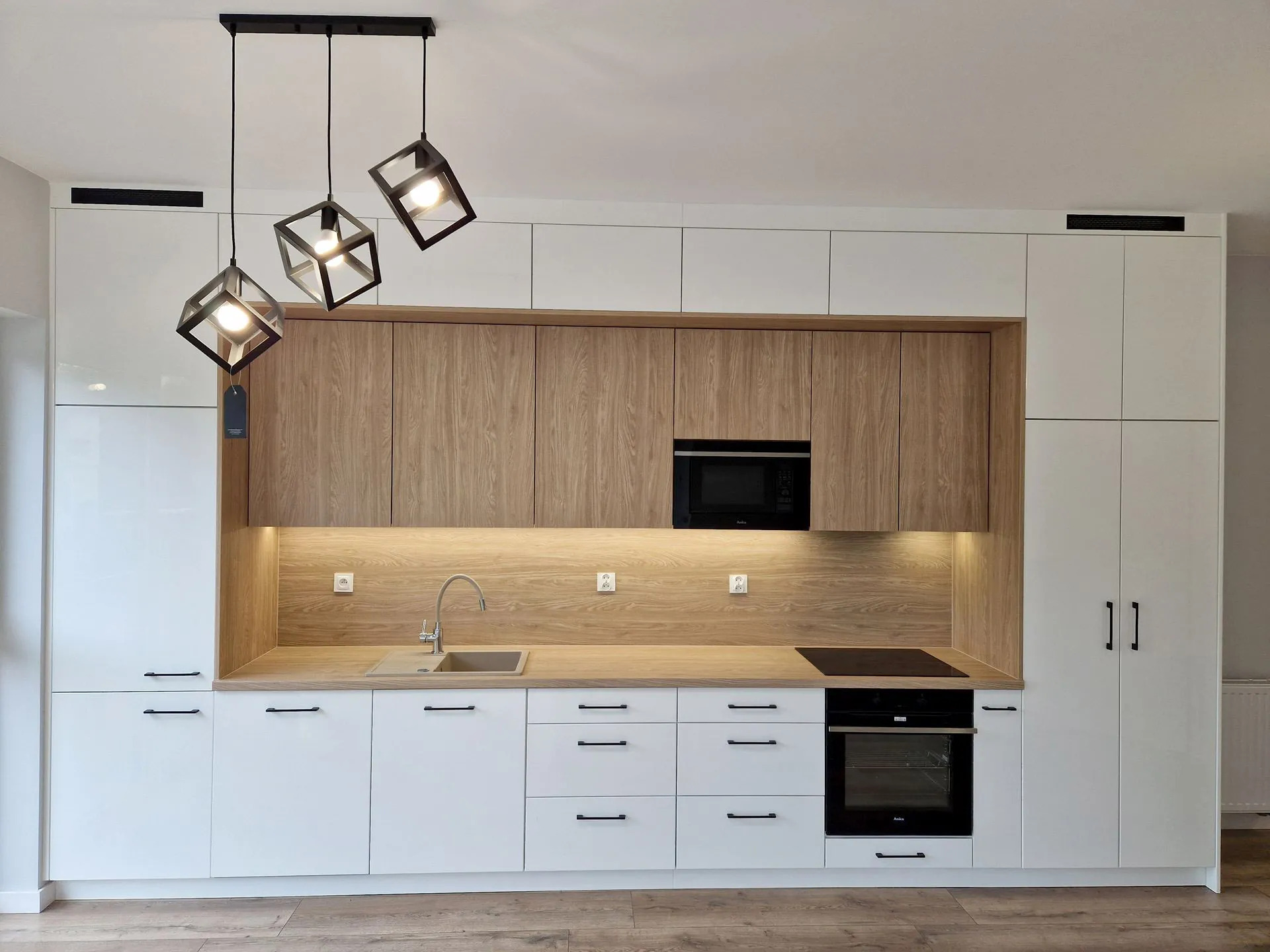 Nowoczesna kuchnia z białymi szafkami dolnymi i drewnopodobnymi górnymi, oświetlona wiszącymi lampami w kształcie sześcianów. Minimalistyczny design z czarnymi uchwytami.