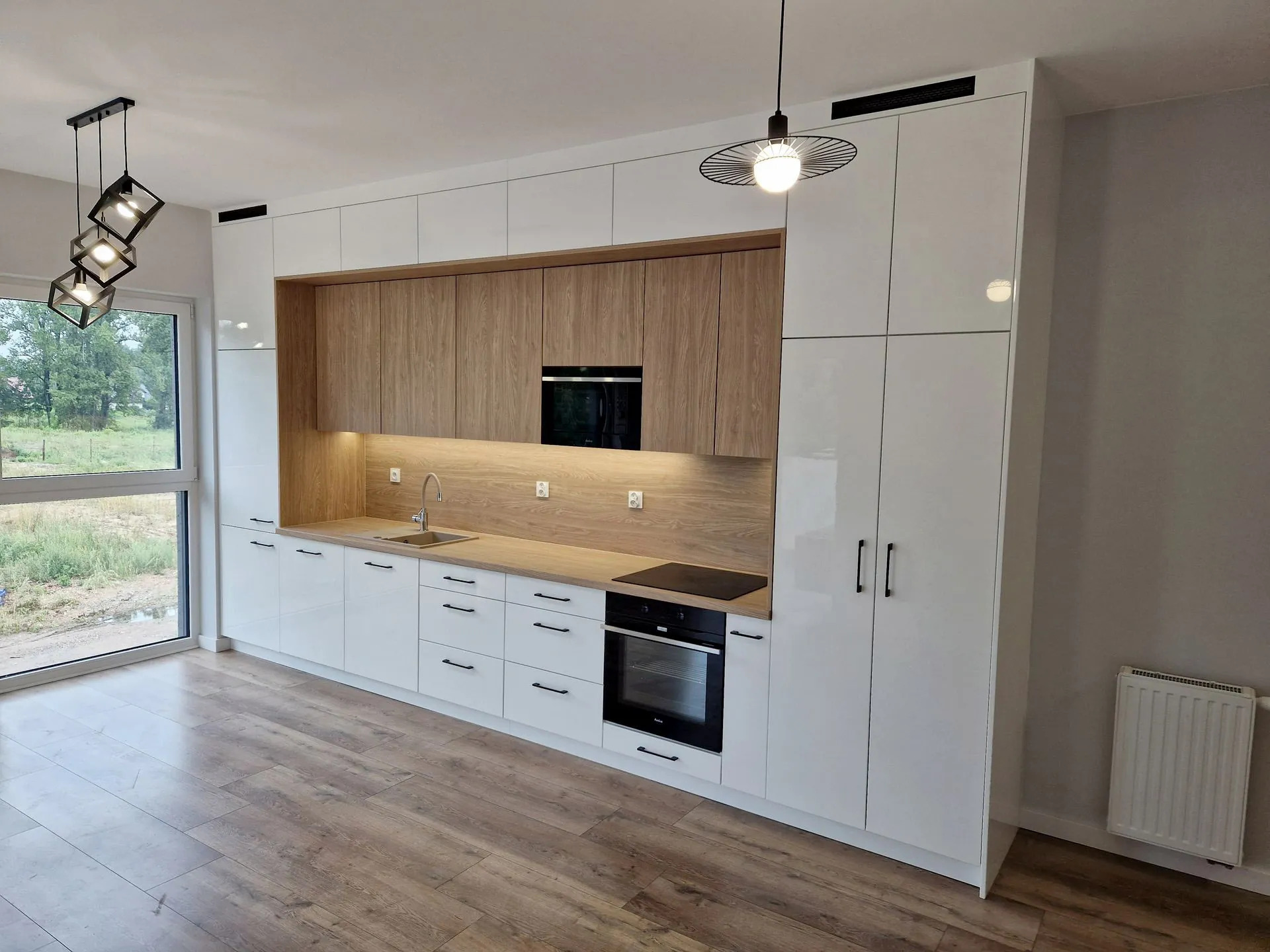 Nowoczesna kuchnia z białymi, lakierowanymi frontami i drewnianymi akcentami w Krakowie. Widoczne oświetlenie podszafkowe i designerskie lampy wiszące.