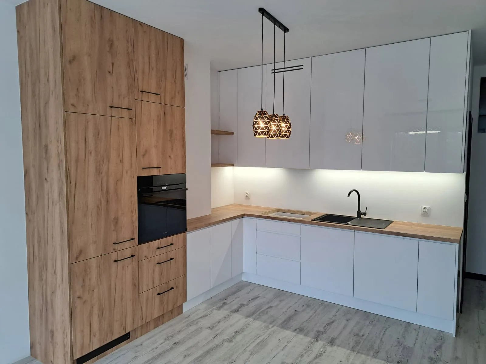 Nowoczesna kuchnia na wymiar z białymi, lakierowanymi frontami i drewnopodobnym wysokim zabudowaniem z piekarnikiem. Wiszące lampy geometryczne.