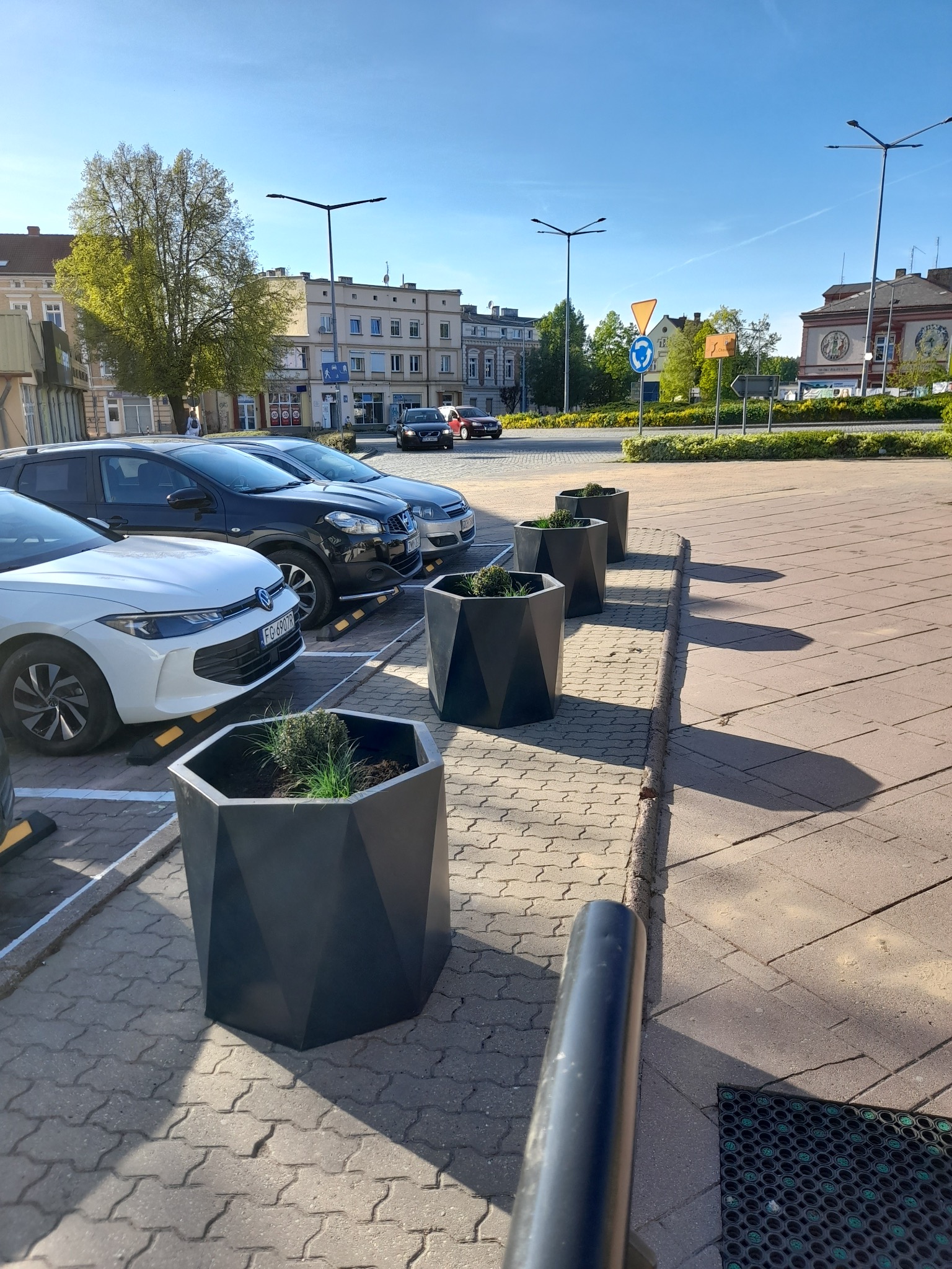 Rząd geometrycznych, grafitowych donic z zielenią, ustawionych wzdłuż chodnika przy parkingu miejskim w słoneczny dzień. Nowoczesny design donic.