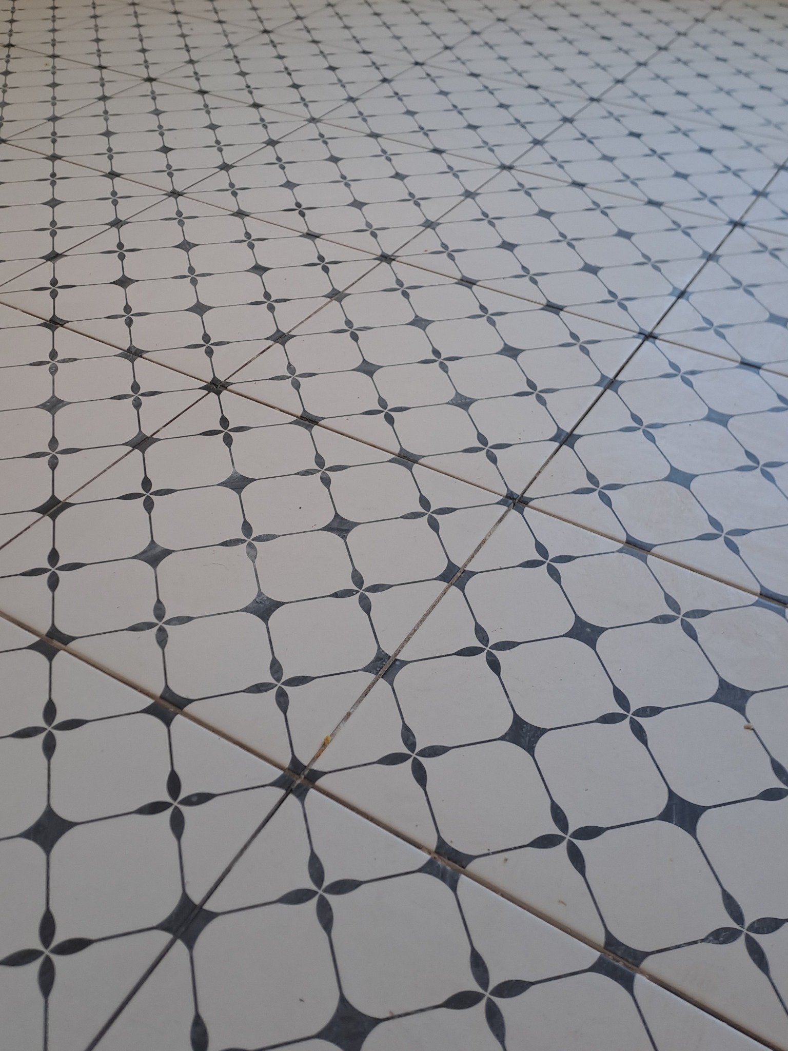 Ułożona podłoga z białych, kwadratowych płytek z ciemnoszarym wzorem w kształcie czterolistnej koniczyny, sfotografowana pod kątem, ukazująca regularność i estetykę wykończenia.