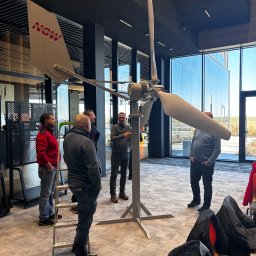 SunTech Jan Karwowski - Wnętrze biura z grupą osób obserwujących model turbiny wiatrowej z napisem 'NOW'. Widoczne duże okna i nowoczesny design.