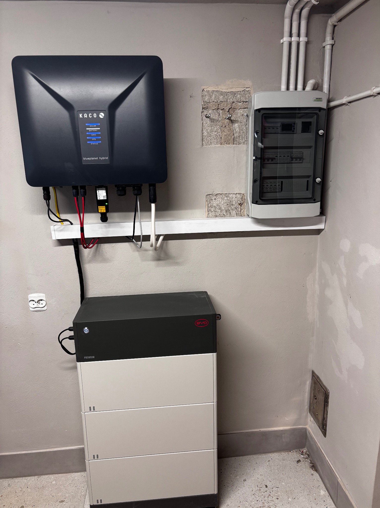 Zainstalowany system fotowoltaiczny z inwerterem Kaco, akumulatorem BYD i rozdzielnicą Noark na ścianie w surowym wnętrzu. Widoczne przewody i gniazdko elektryczne.