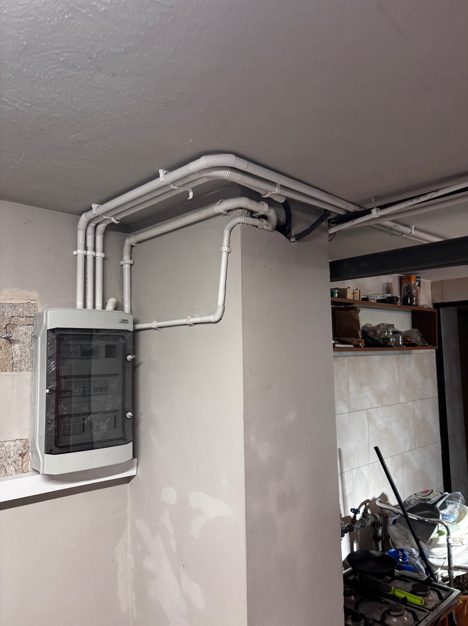 Rozdzielnia elektryczna natynkowa z białymi rurami instalacyjnymi pod sufitem w pomieszczeniu z elementami wykończenia kuchennego w tle.