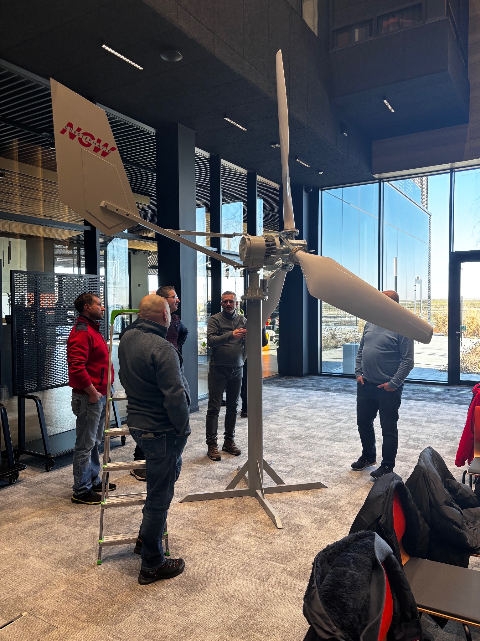 Wnętrze biura z grupą osób obserwujących model turbiny wiatrowej z napisem 'NOW'. Widoczne duże okna i nowoczesny design.