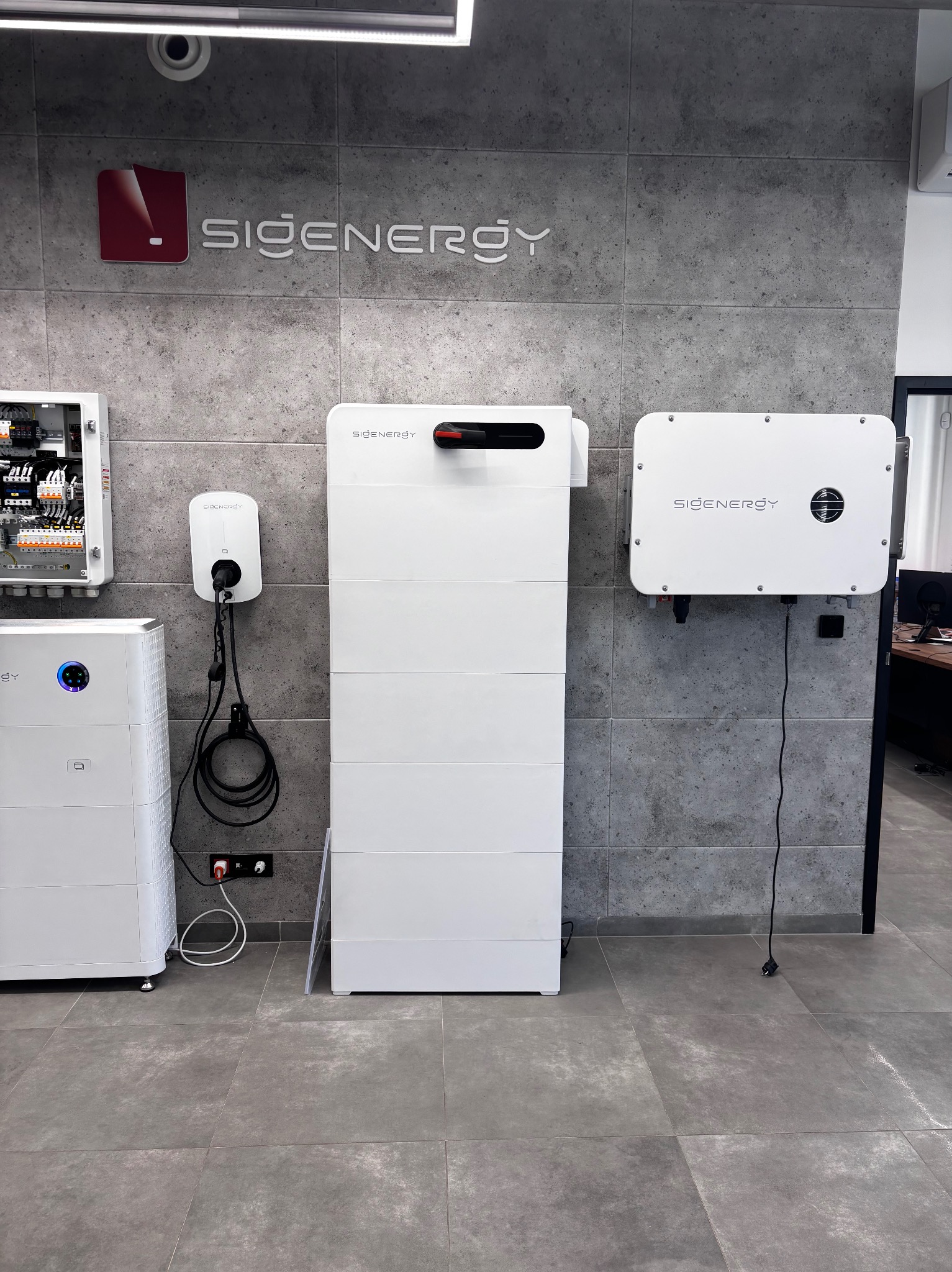 Kompletny system fotowoltaiczny z magazynem energii i ładowarką EV, zamontowany na szarej ścianie z płytek. Widoczne kable i elementy instalacji.