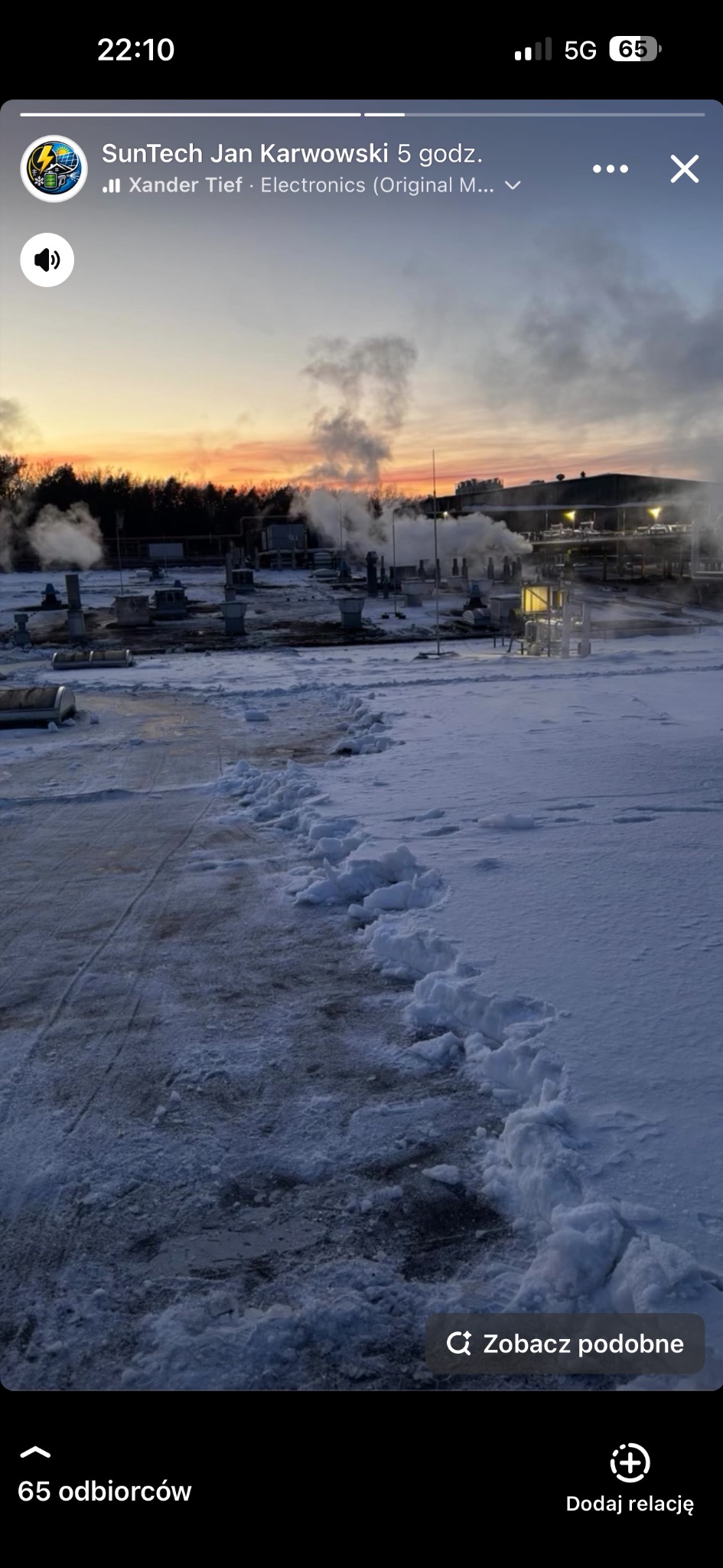 Śnieżny teren przemysłowy o zmierzchu. Widoczne instalacje, rury i budynki w tle. Ślady na śniegu prowadzą w stronę instalacji. Dym unosi się w powietrzu.