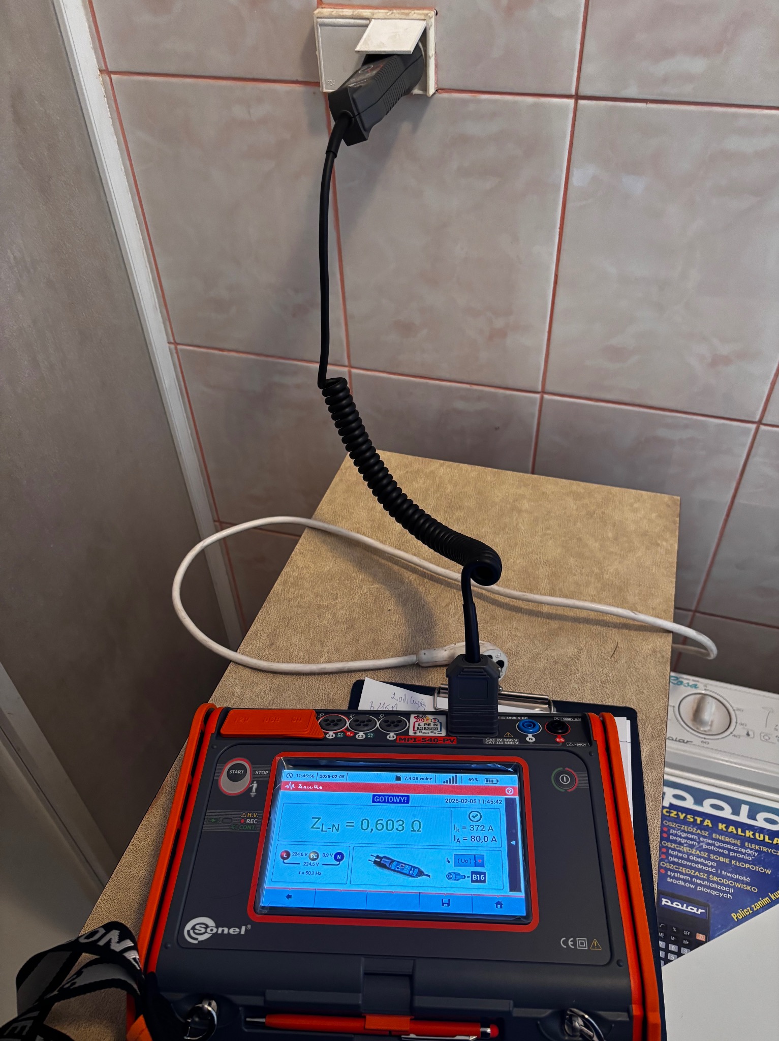 Miernik Sonel MPI-540-PV podłączony do gniazdka elektrycznego podczas pomiarów. Widoczne parametry na ekranie urządzenia i spiralny kabel zasilający.