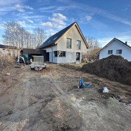 ROM-BRUK - Przygotowanie terenu pod budowę domu jednorodzinnego. Widoczny mini-koparka, przyczepa, hałda ziemi i częściowo wykończone budynki w tle, błękitne niebo.