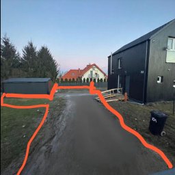 ROM-BRUK - Utwardzona droga dojazdowa do nowoczesnego, czarnego domu z numerem 10A, widok z perspektywy. W tle drugi dom z czerwonym dachem i zielone drzewa.