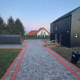 ROM-BRUK - Ułożona kostka brukowa w odcieniach szarości z czerwoną obwódką tworząca ścieżkę do nowoczesnego domu z czarną elewacją i drewnianym podjazdem dla niepełnosprawnych.