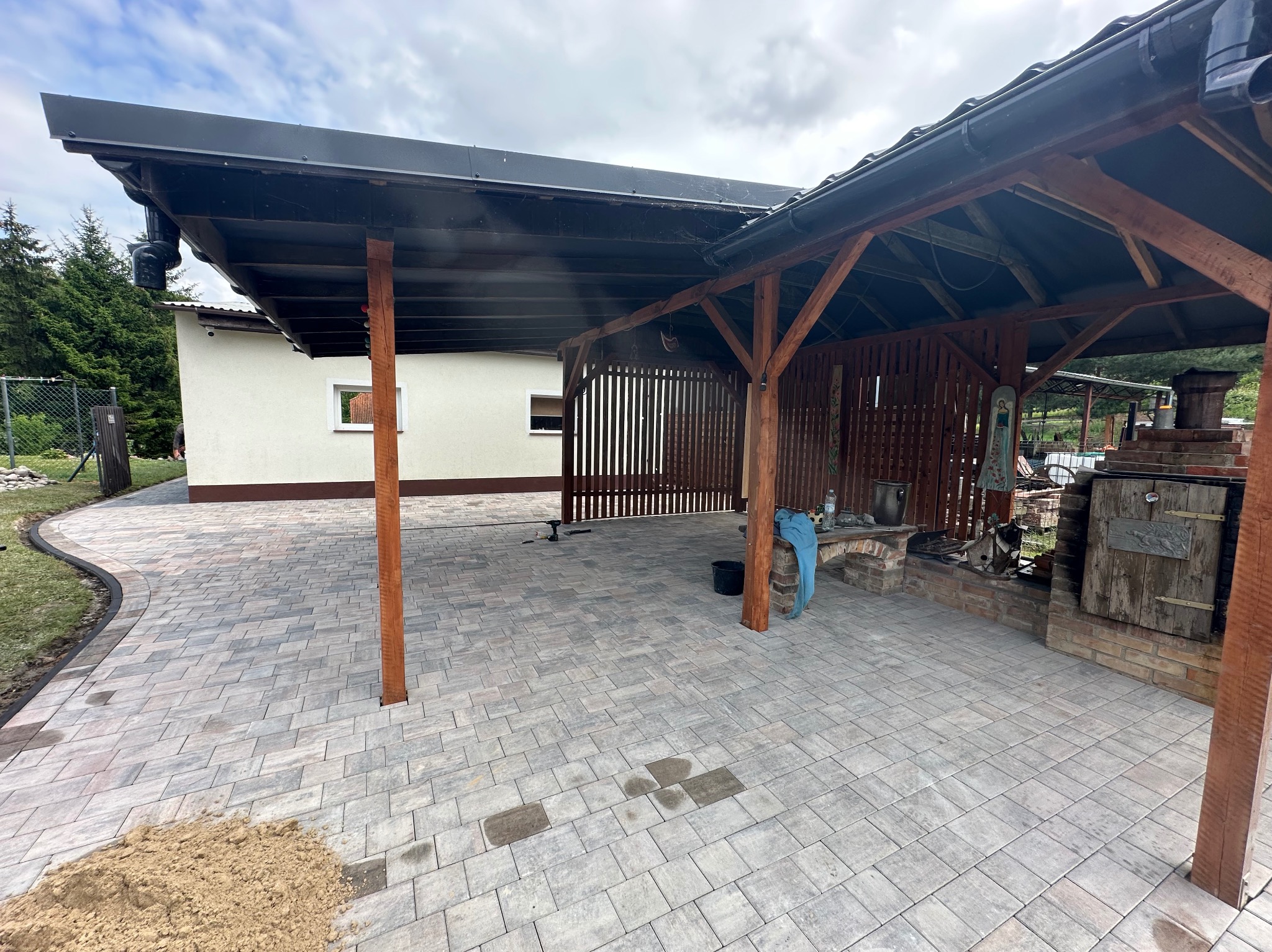 Ułożona kostka brukowa w odcieniach szarości tworzy wzór na podjeździe z drewnianą konstrukcją zadaszenia. Widoczny fragment domu i zieleni w tle.