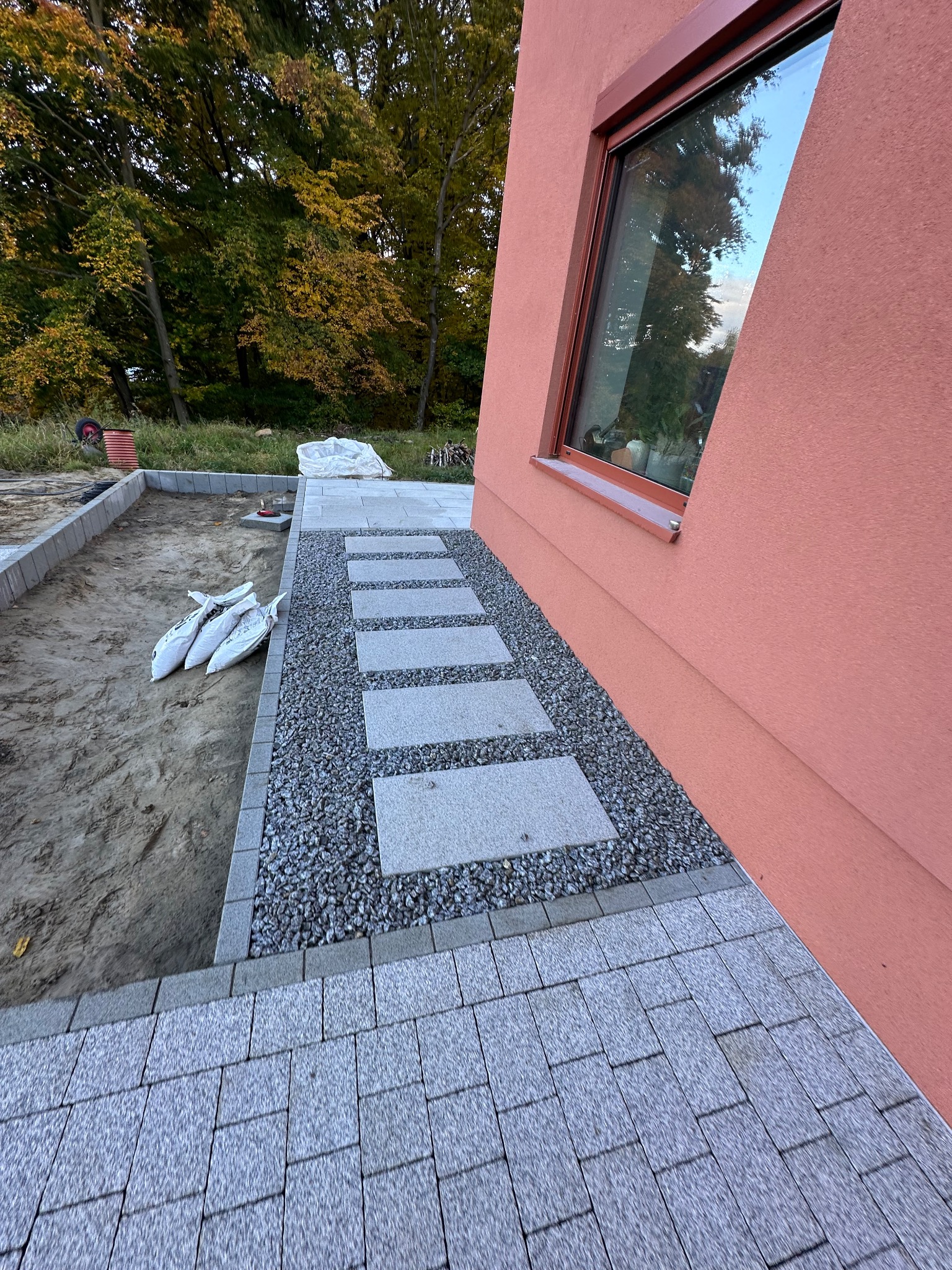 Ścieżka z płyt i grysu przy budynku z oknem. Fragment ułożonej kostki brukowej i przygotowany teren. Worki z materiałem budowlanym w tle.