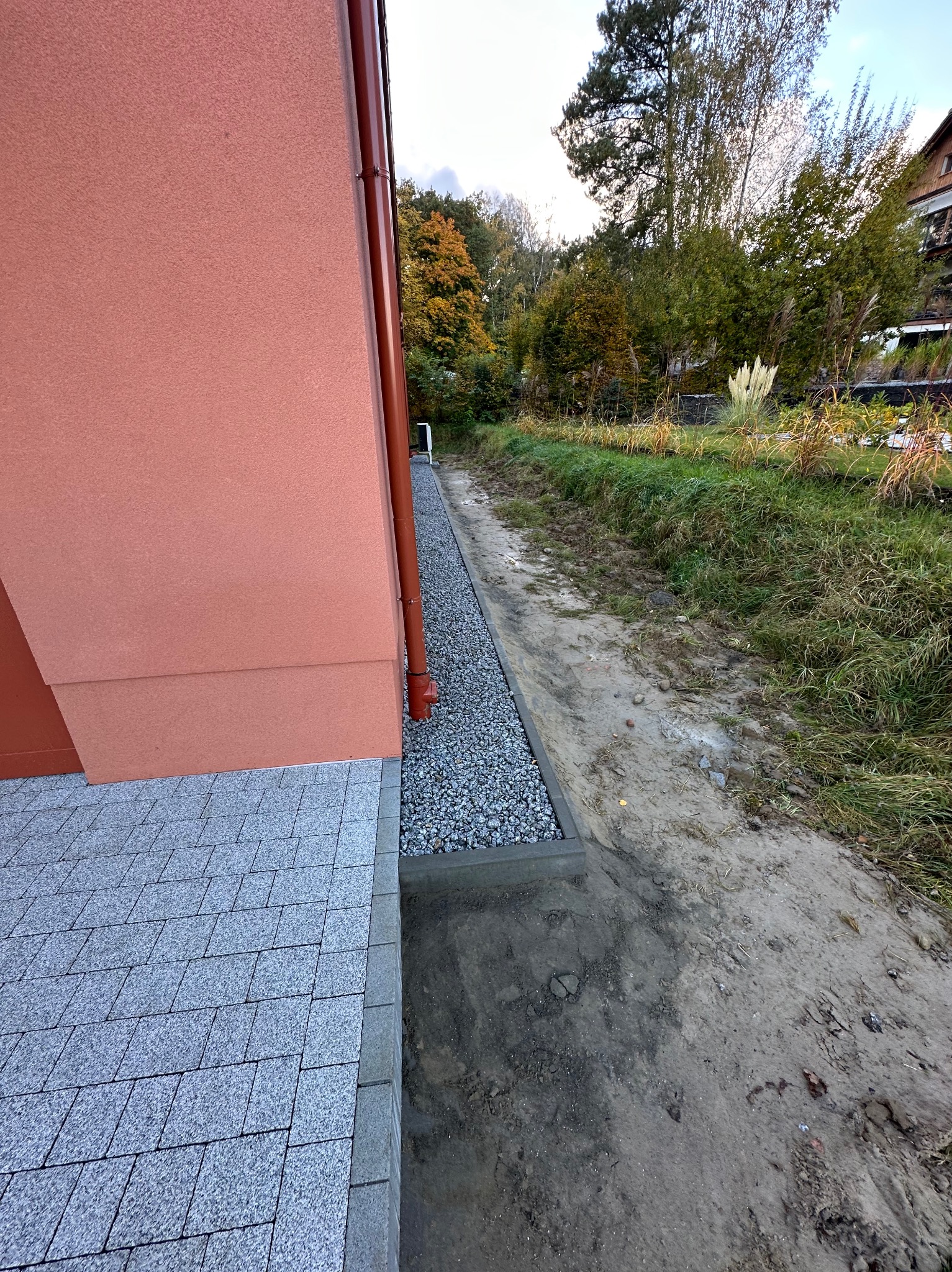 Szara kostka brukowa ułożona przy ścianie budynku z brązową rynną, obok wąski pas wysypany drobnym żwirem, dalej ziemia z roślinnością.