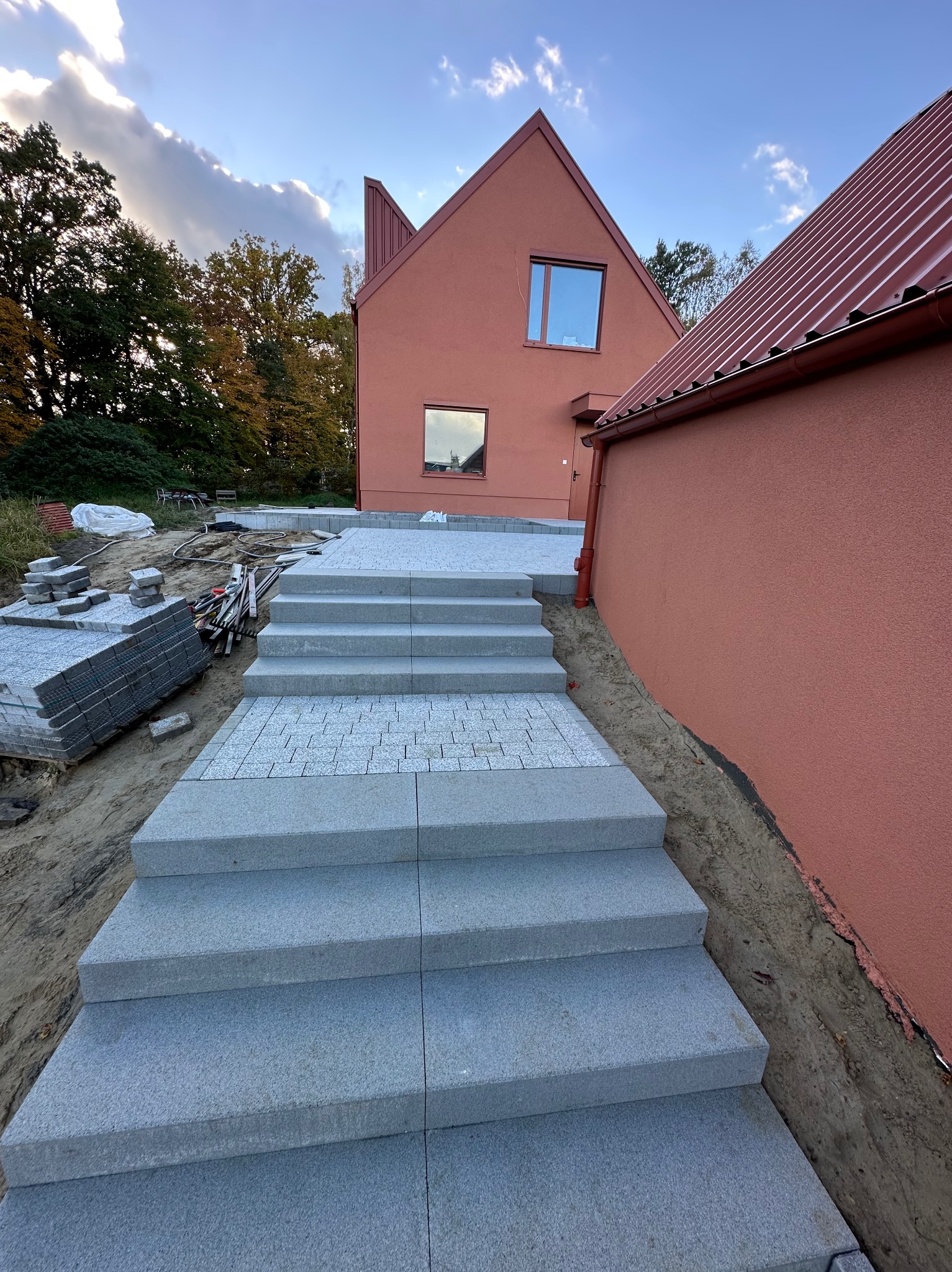 Granitowe schody prowadzące do domu o ceglanej elewacji, częściowo wykończone kostką. Widoczne zapasy materiału i narzędzia brukarskie. Praca w toku, etap układania.