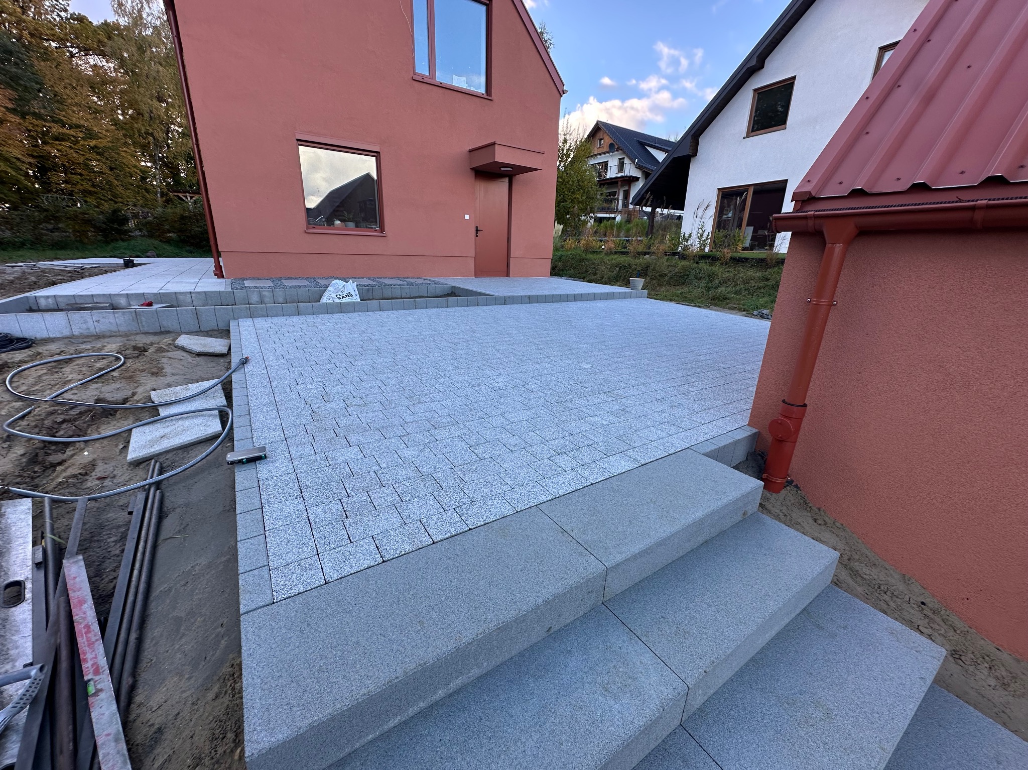Ułożona szara kostka granitowa tworząca taras i schody przed domem z czerwoną elewacją. Widoczne narzędzia i worki z materiałem budowlanym.
