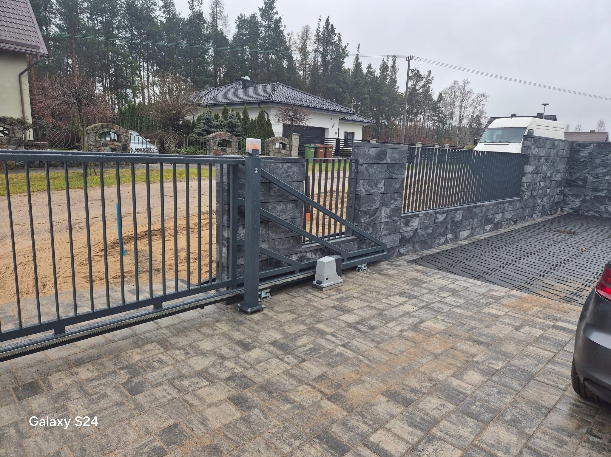 Nowoczesna, przesuwna brama wjazdowa z szarymi przęsłami i automatyką, osadzona na kostce brukowej w odcieniach beżu i szarości. W tle dom jednorodzinny i zieleń.