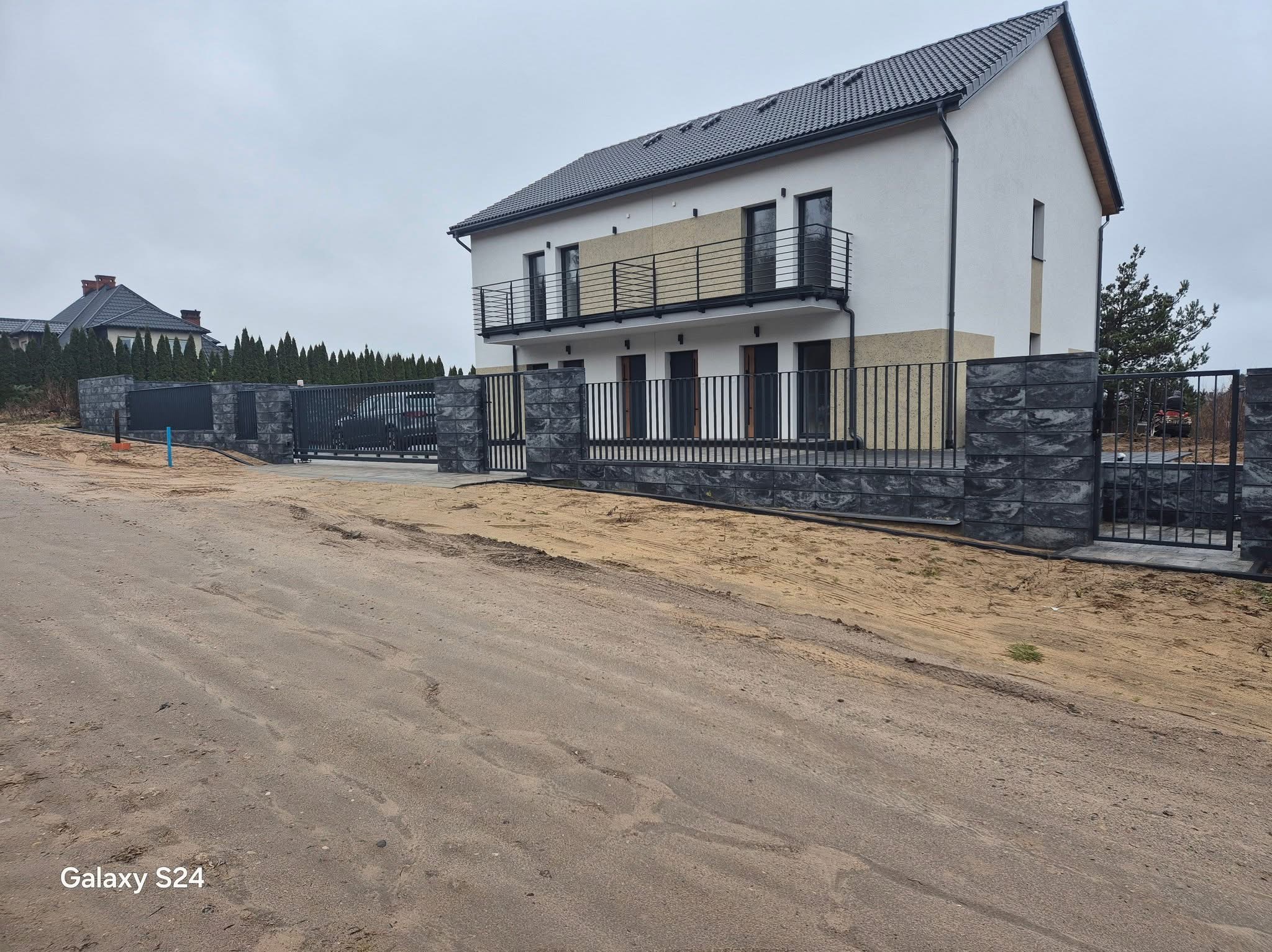 Nowoczesny dom z grafitowym ogrodzeniem z elementami betonowymi. Metalowe przęsła i brama w kolorze antracytu. Surowy teren przed posesją.