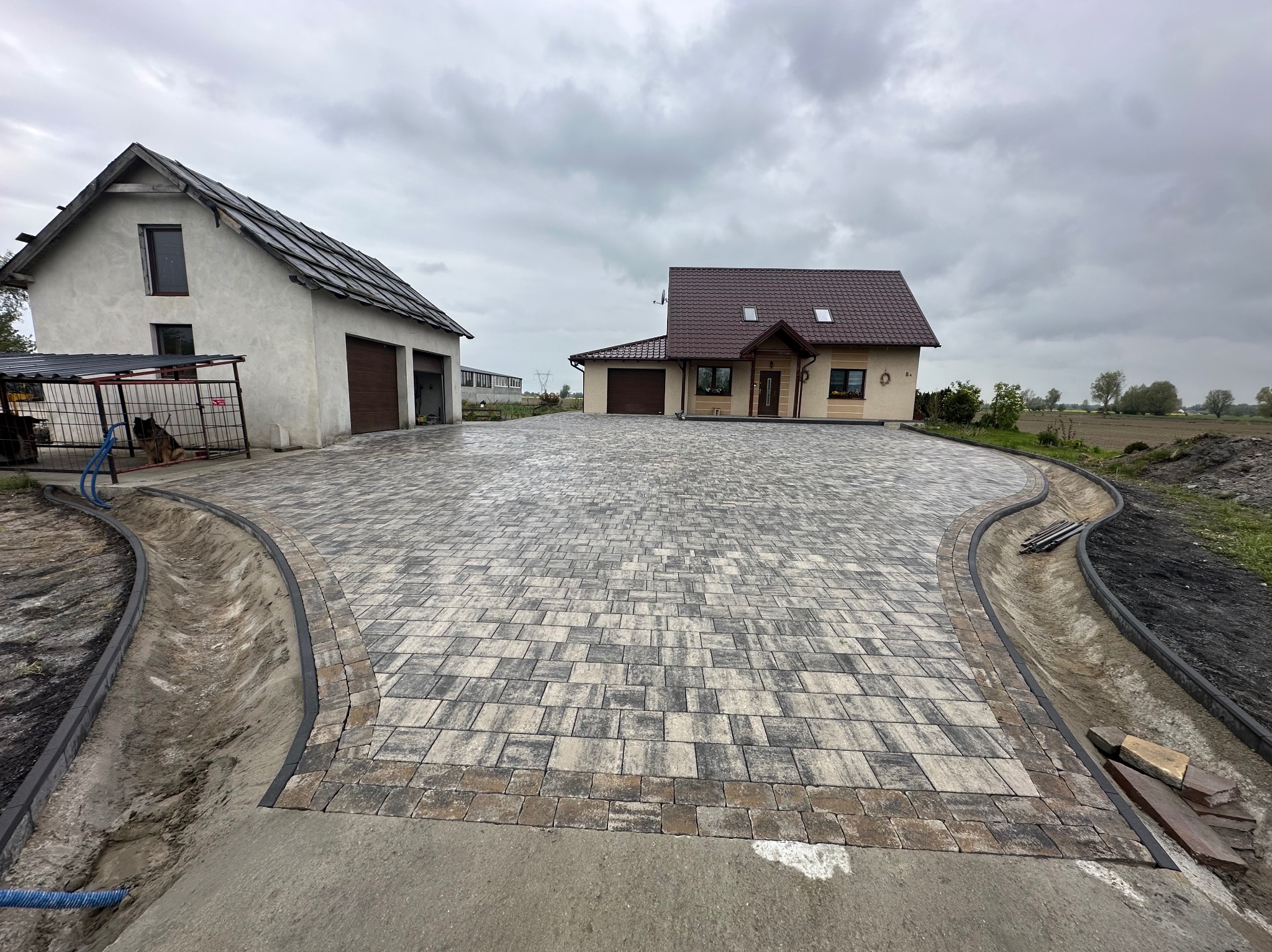 Szeroki podjazd z kostki brukowej prowadzący do domu i garażu, obrzeża z krawężników, widok na budynki w tle, pochmurne niebo.