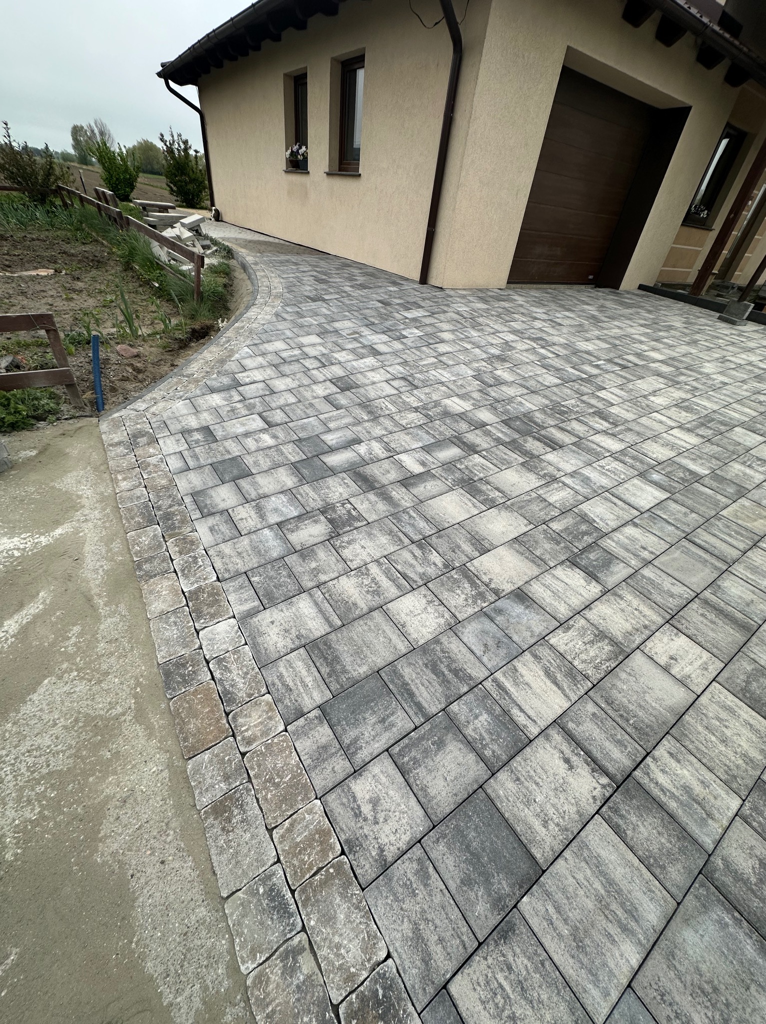 Nowoczesny podjazd z szarej kostki brukowej prowadzący do domu z beżową elewacją i brązową bramą garażową. Widoczne obrzeża i fragment ogrodu.