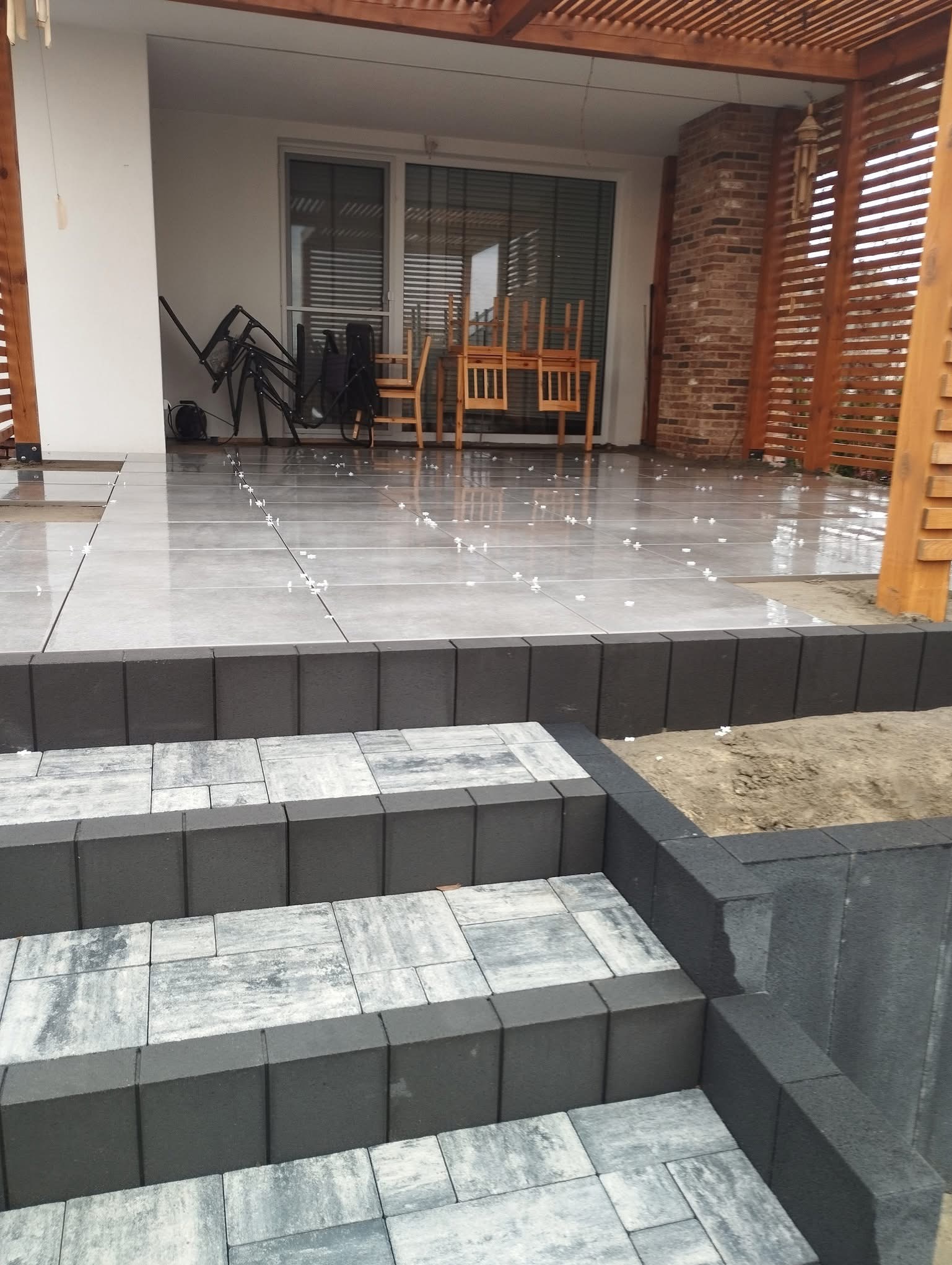 Schody z kostki brukowej prowadzące na taras z szarych płytek. Widoczne białe krzyżyki dystansowe. W tle zadaszenie tarasu i meble ogrodowe.