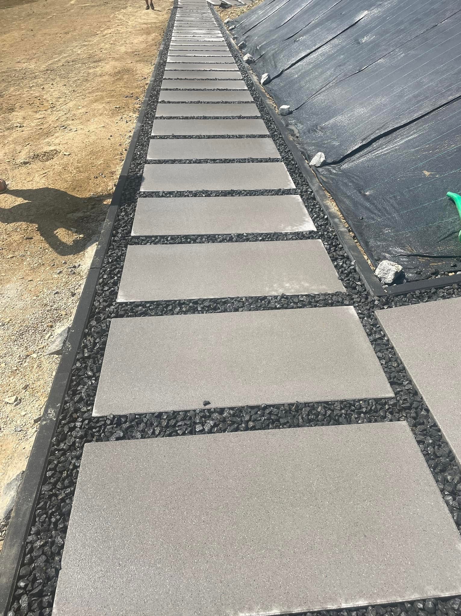 Nowoczesna ścieżka z szarych płyt, oddzielonych czarnym grysem, prowadząca wzdłuż skarpy zabezpieczonej czarną geowłókniną. Minimalistyczny design.