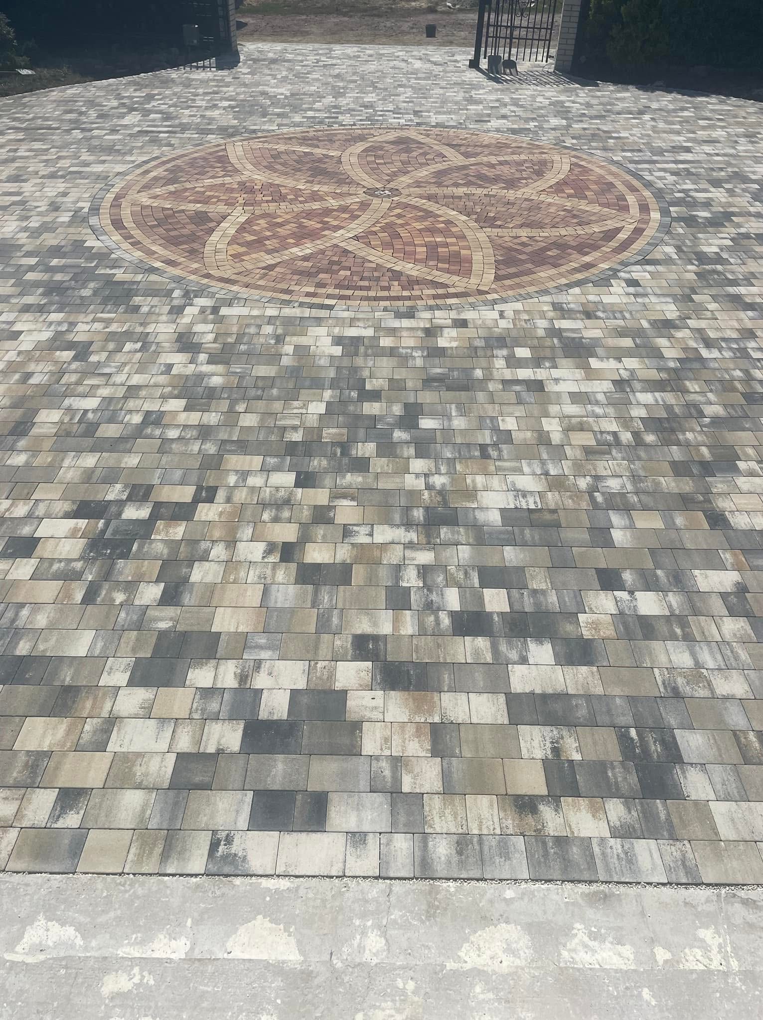 Oryginalny wzór z kostki brukowej w Zblewie. Okrągły ornament z brązowej kostki na tle szaro-beżowej nawierzchni. Widok na bramę w tle. Estetyczne wykończenie.