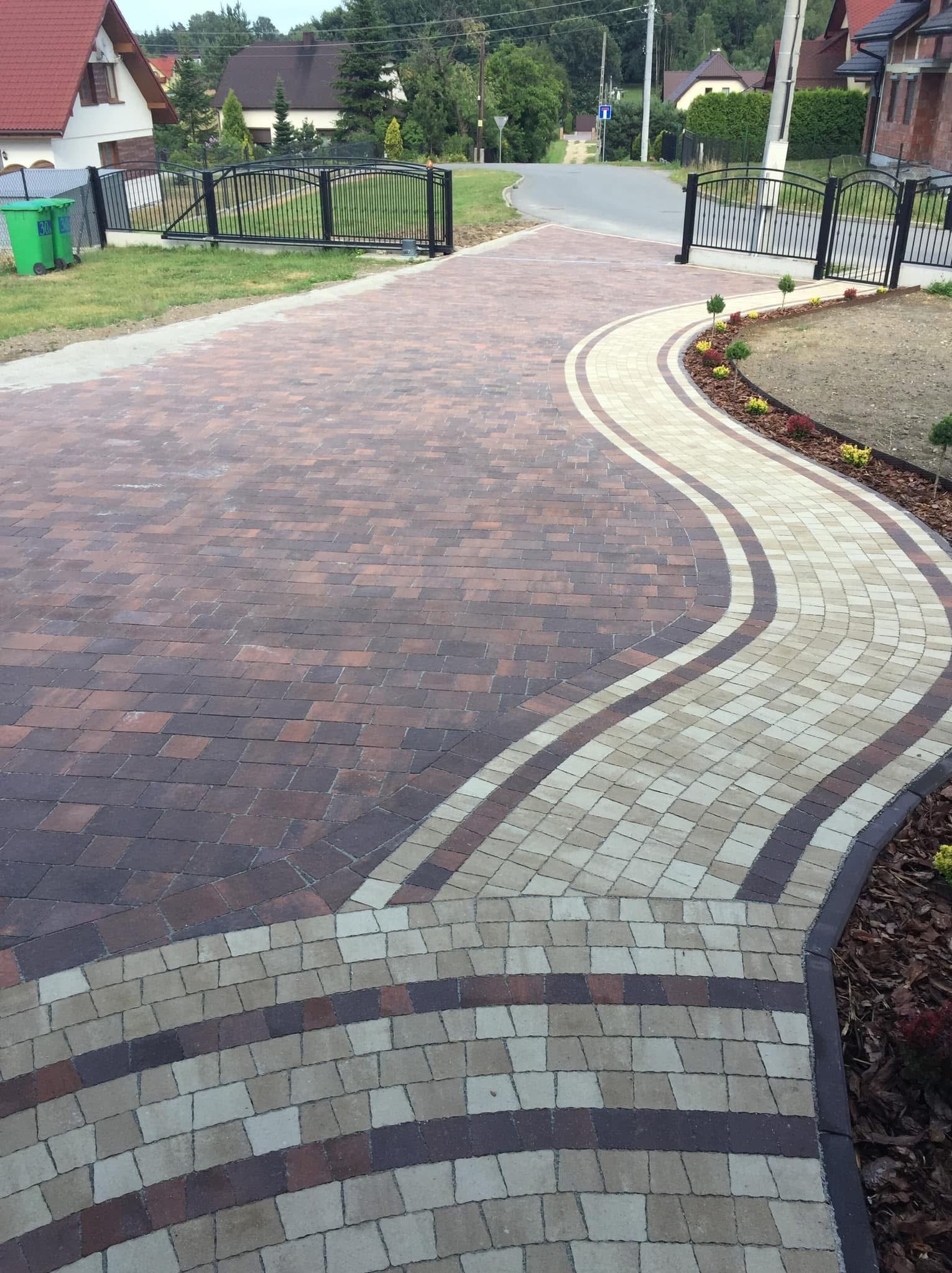 Wzorzysty podjazd z kostki brukowej w odcieniach brązu i beżu, wijący się łukiem przez posesję. Widoczne ogrodzenie z bramą i zadbana zieleń.