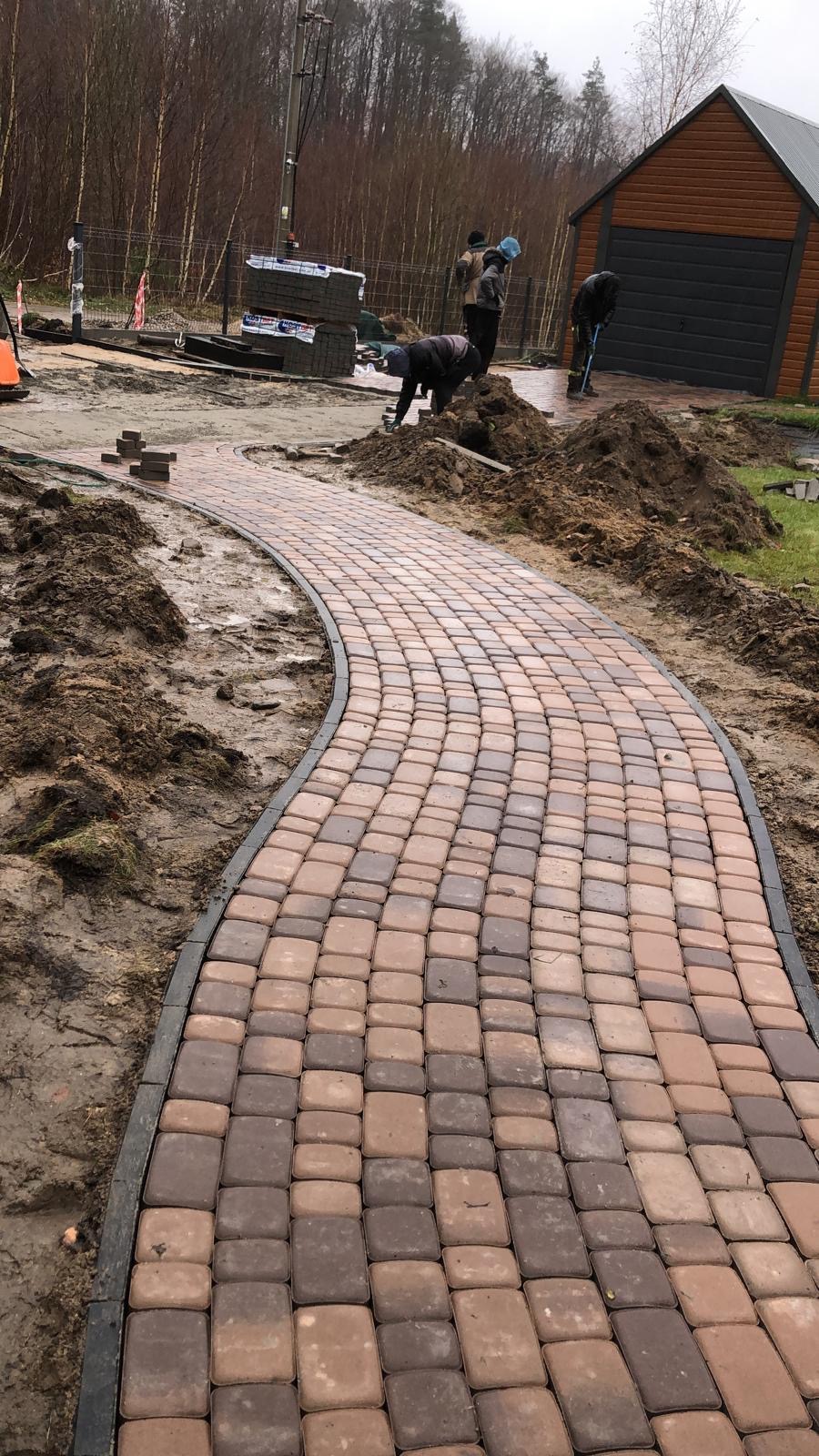 Układanie krętej ścieżki z kostki brukowej w odcieniach brązu i beżu. W tle sterta kostki, pracownicy i budynek gospodarczy. Widok na częściowo ukończony projekt.