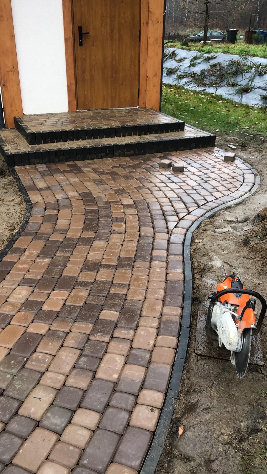 Kaskadowe schody i ścieżka z kostki brukowej w odcieniach brązu, wykończona czarnym obrzeżem. W tle drzwi wejściowe i fragment ogrodu. Obok leży piła do cięcia kostki.