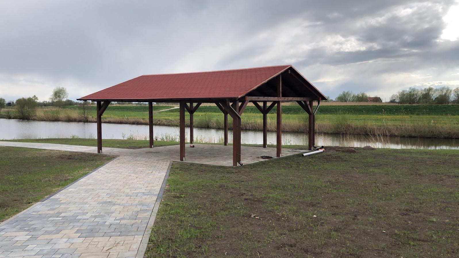 Altana z czerwoną dachówką nad brzegiem jeziora, z brukowanym chodnikiem prowadzącym do niej. Widok na spokojną wodę i zielone brzegi pod pochmurnym niebem.