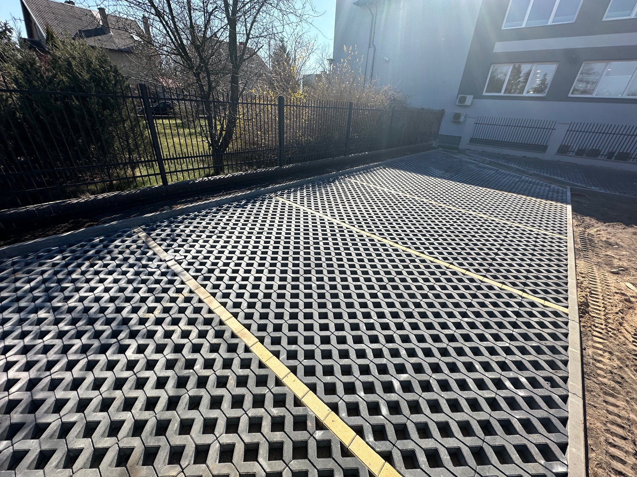 Szara, ażurowa nawierzchnia z kostki, z żółtymi liniami oddzielającymi miejsca parkingowe, na tle budynku i ogrodzenia. Widoczny fragment terenu przed ułożeniem nawierzchni.