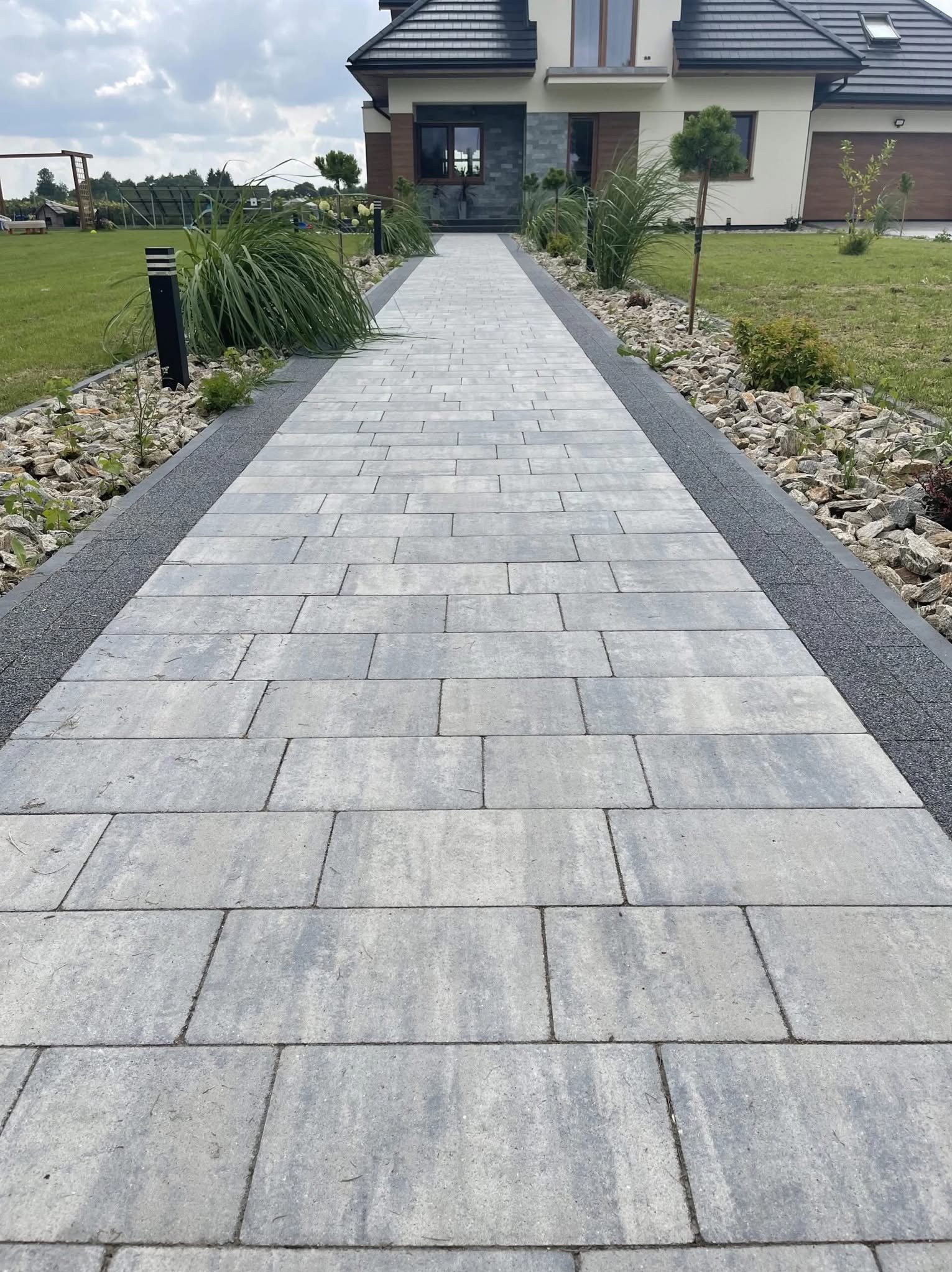 Długa, prosta ścieżka z szarej kostki brukowej prowadzi do domu. Po bokach ścieżki ozdobne kamienie i roślinność. Widok na dom z ciemnym dachem i jasną elewacją.