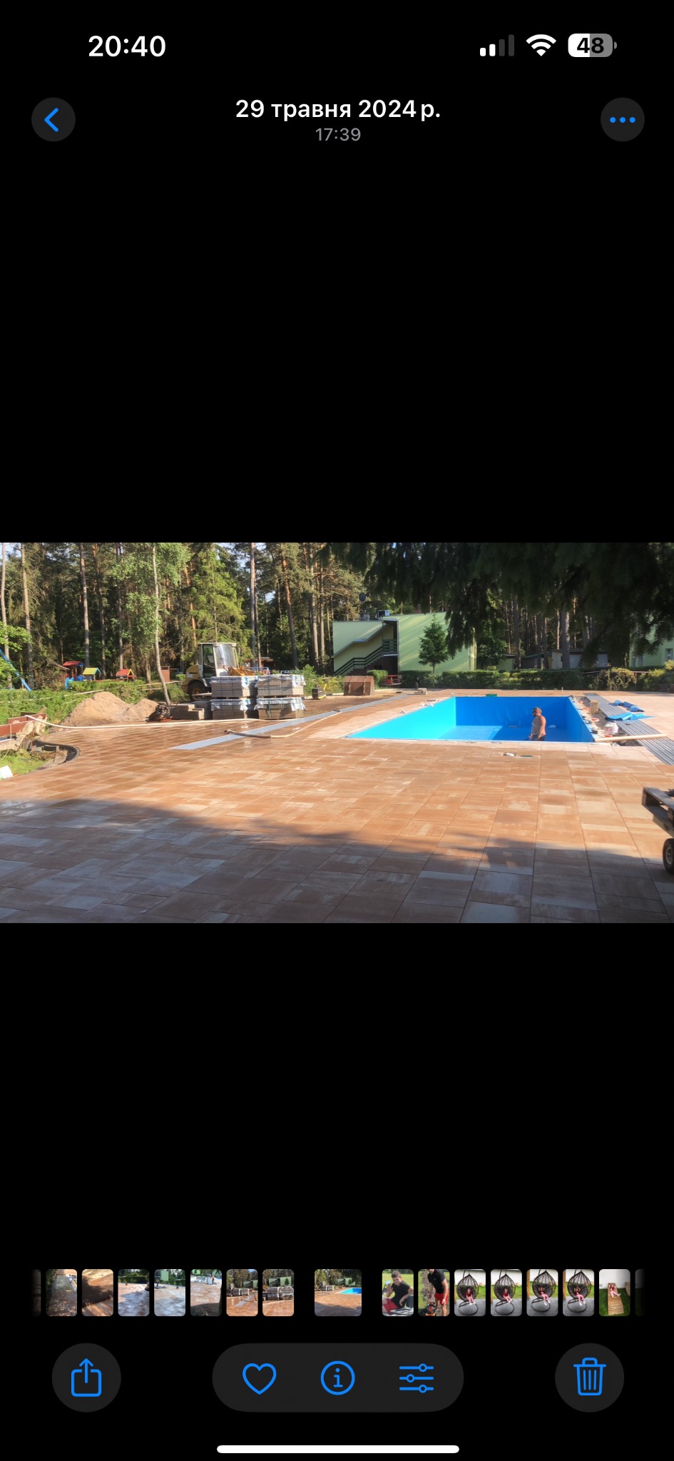 Brązowa kostka brukowa wokół basenu w ogrodzie, w tle las i budynek. Częściowo ułożona nawierzchnia, widoczne palety z kostką i maszyna budowlana.