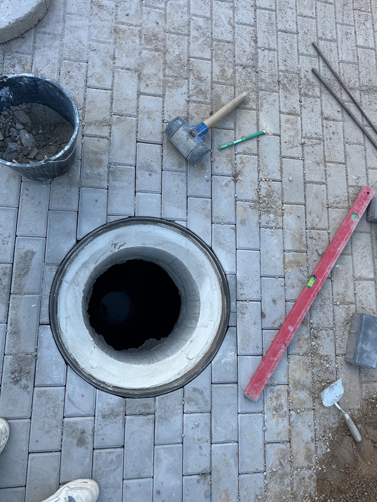Szara kostka brukowa ułożona wokół betonowego kręgu studni. Widoczne narzędzia: poziomica, kielnia, młotek brukarski, wiadro z zaprawą. Fragment buta w lewym dolnym rogu.