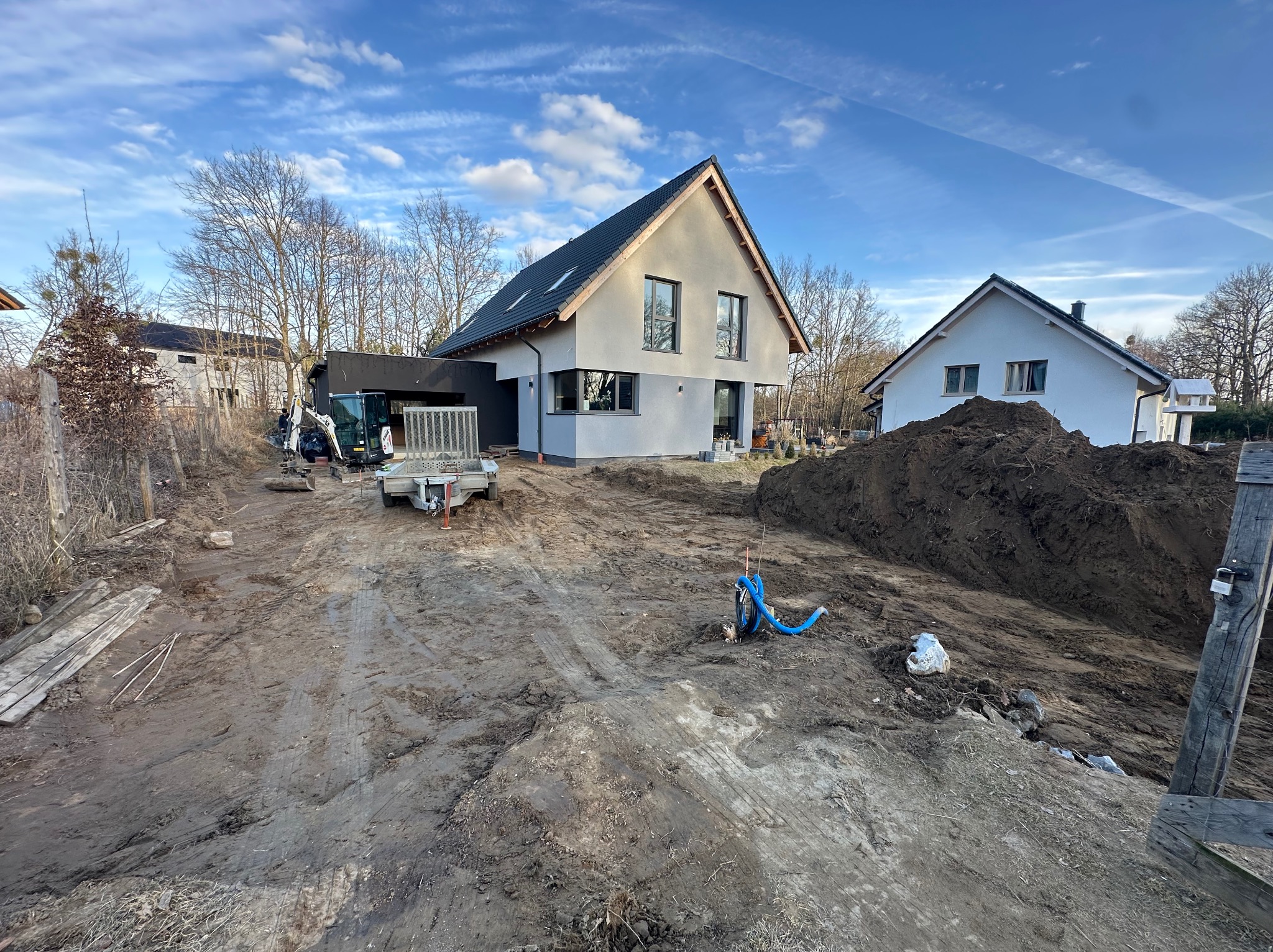 Przygotowanie terenu pod budowę domu jednorodzinnego. Widoczny mini-koparka, przyczepa, hałda ziemi i częściowo wykończone budynki w tle, błękitne niebo.