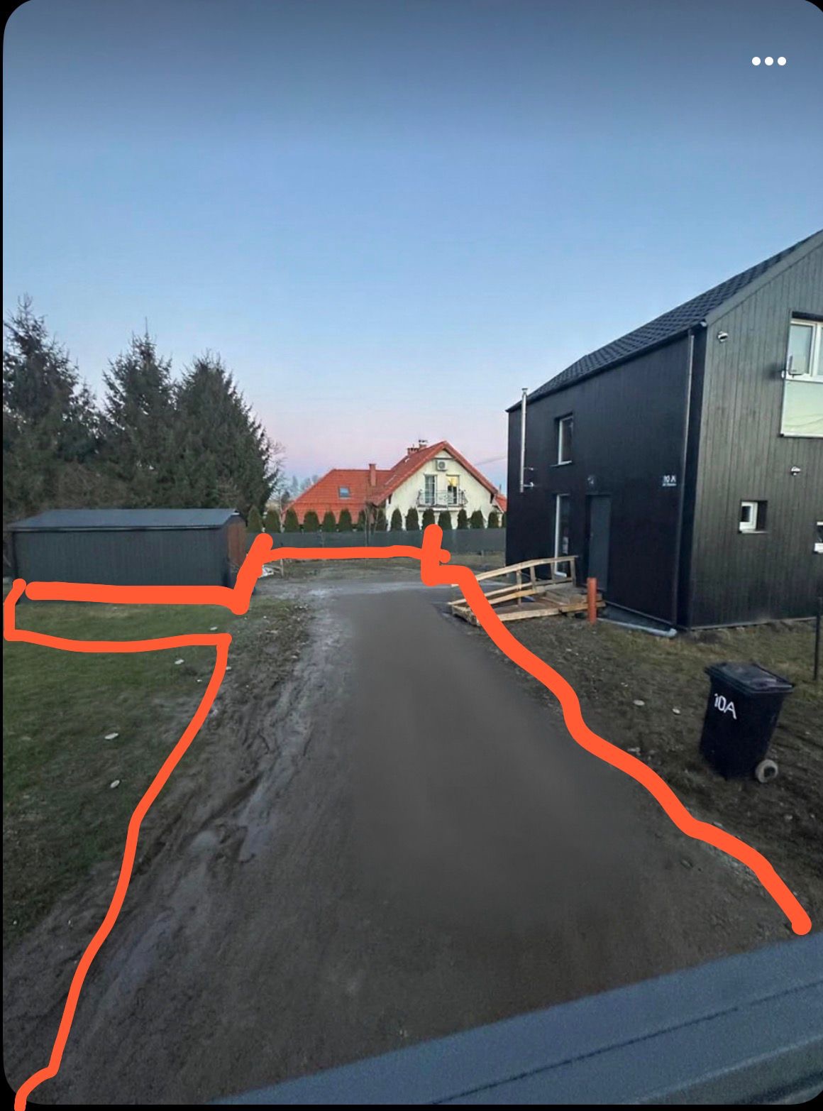 Utwardzona droga dojazdowa do nowoczesnego, czarnego domu z numerem 10A, widok z perspektywy. W tle drugi dom z czerwonym dachem i zielone drzewa.