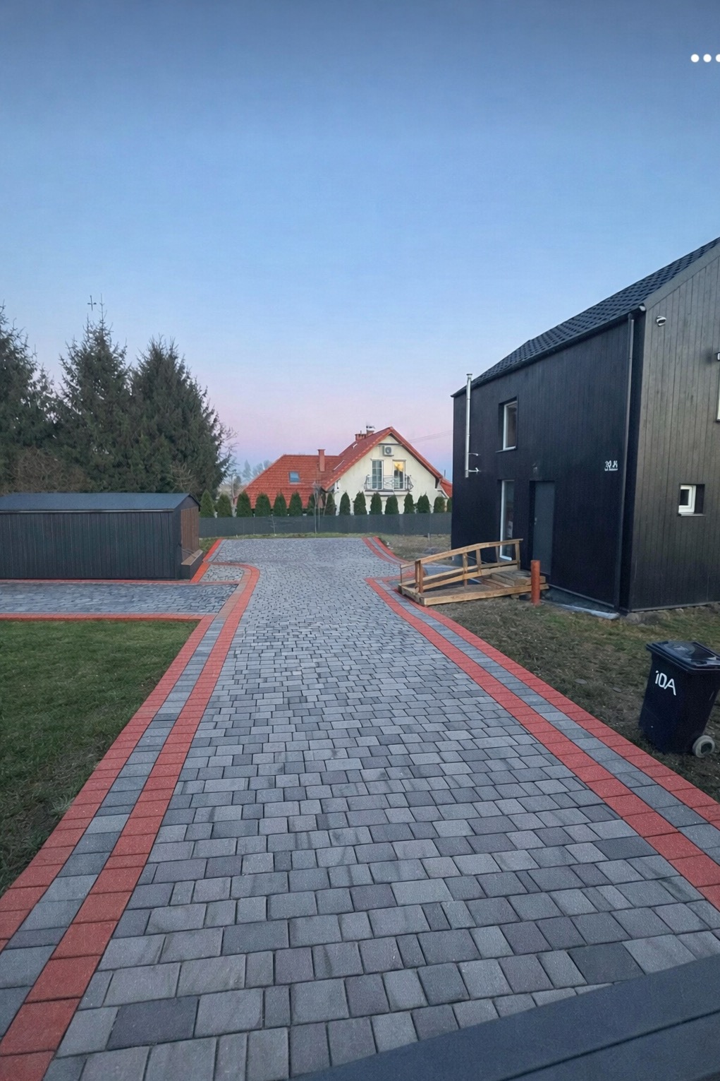 Ułożona kostka brukowa w odcieniach szarości z czerwoną obwódką tworząca ścieżkę do nowoczesnego domu z czarną elewacją i drewnianym podjazdem dla niepełnosprawnych.