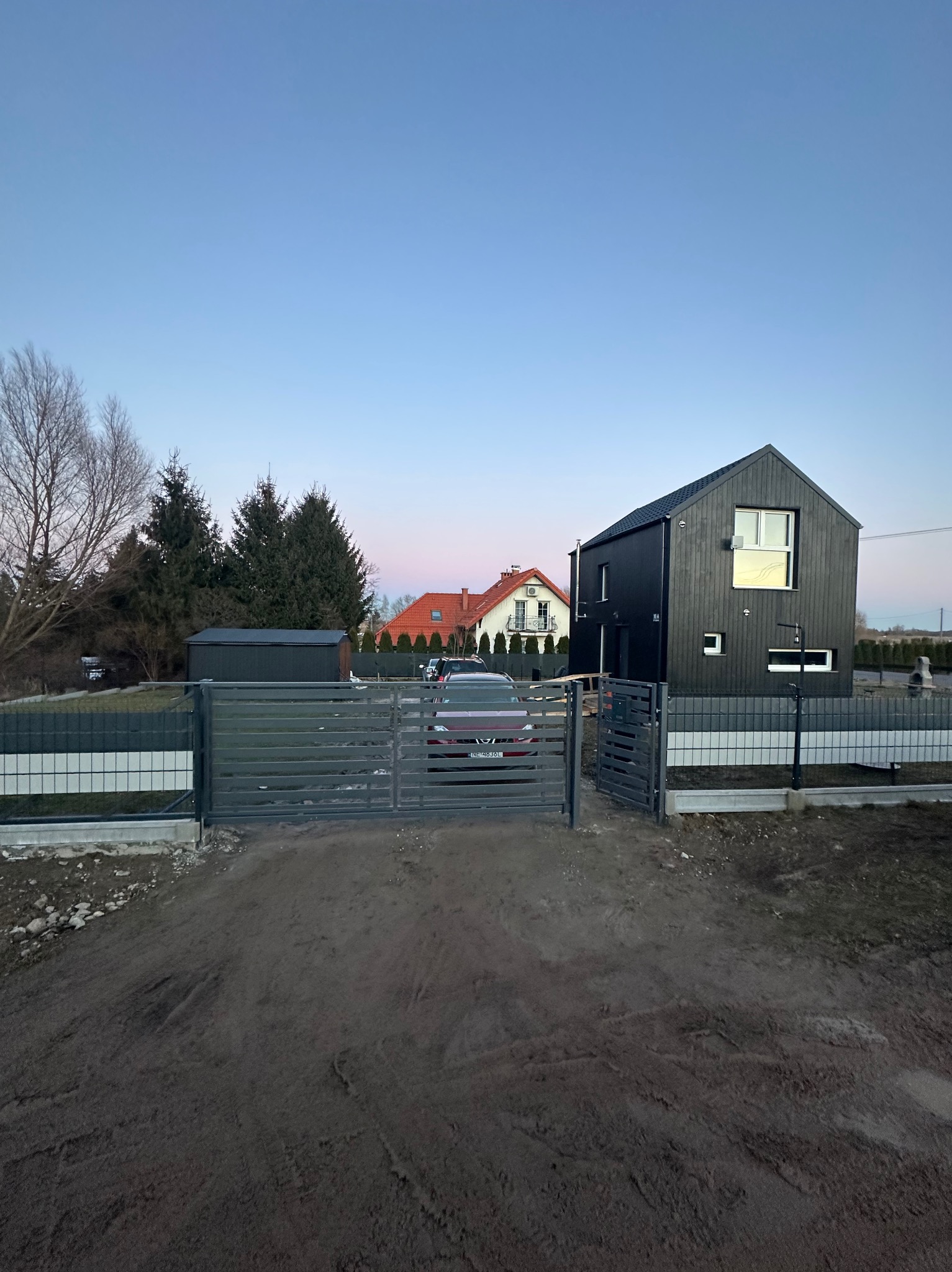 Nowoczesne, poziome ogrodzenie panelowe w kolorze grafitowym z otwartą bramą wjazdową na tle domu o ciemnej elewacji i nieba o zmierzchu.