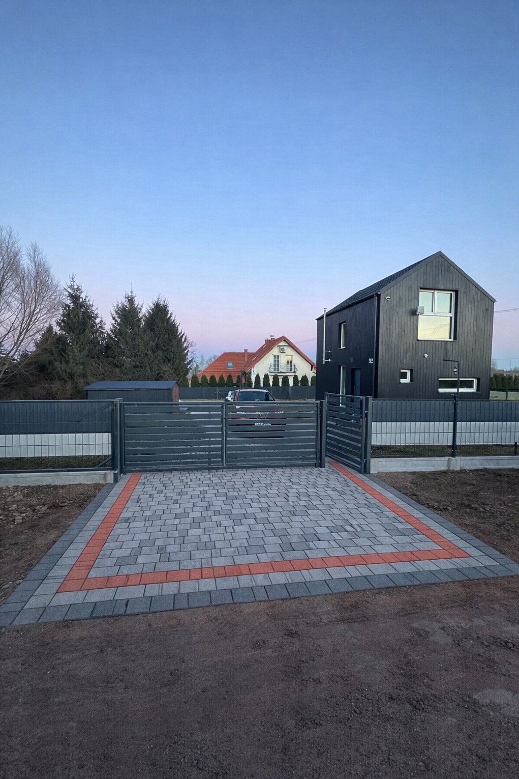 Nowoczesny podjazd z kostki brukowej w odcieniach szarości i czerwieni, prowadzący do posesji z czarnym domem i szarym ogrodzeniem panelowym.