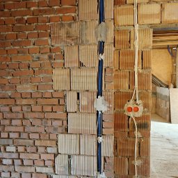 Złota Faza Michał Wacławik Instalacje elektryczne - Murowana ściana z widocznymi przewodami w peszlu i puszkami instalacyjnymi. Surowy stan, instalacja w trakcie montażu, widoczne elementy konstrukcji dachu.