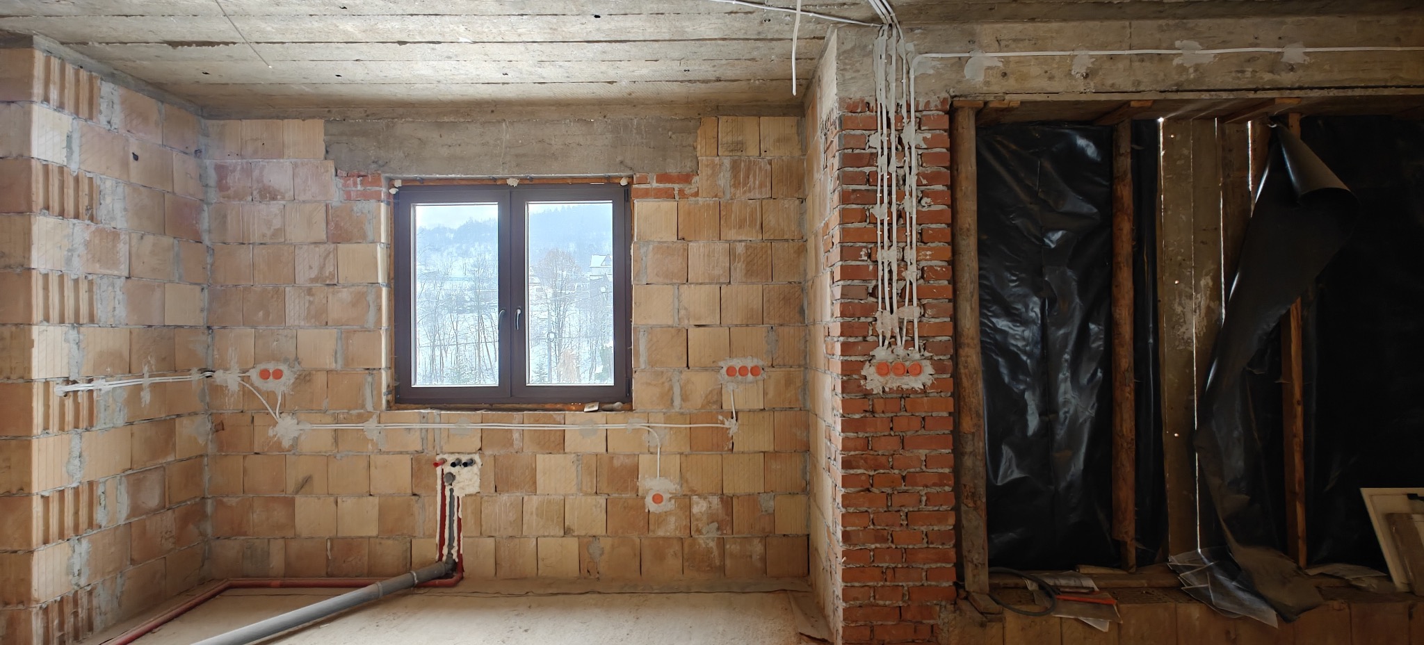 Surowy stan: Instalacja elektryczna w ścianach z pustaków ceramicznych, widoczne kable i puszki. Okno z widokiem na miasto w tle. Prace instalacyjne w trakcie.